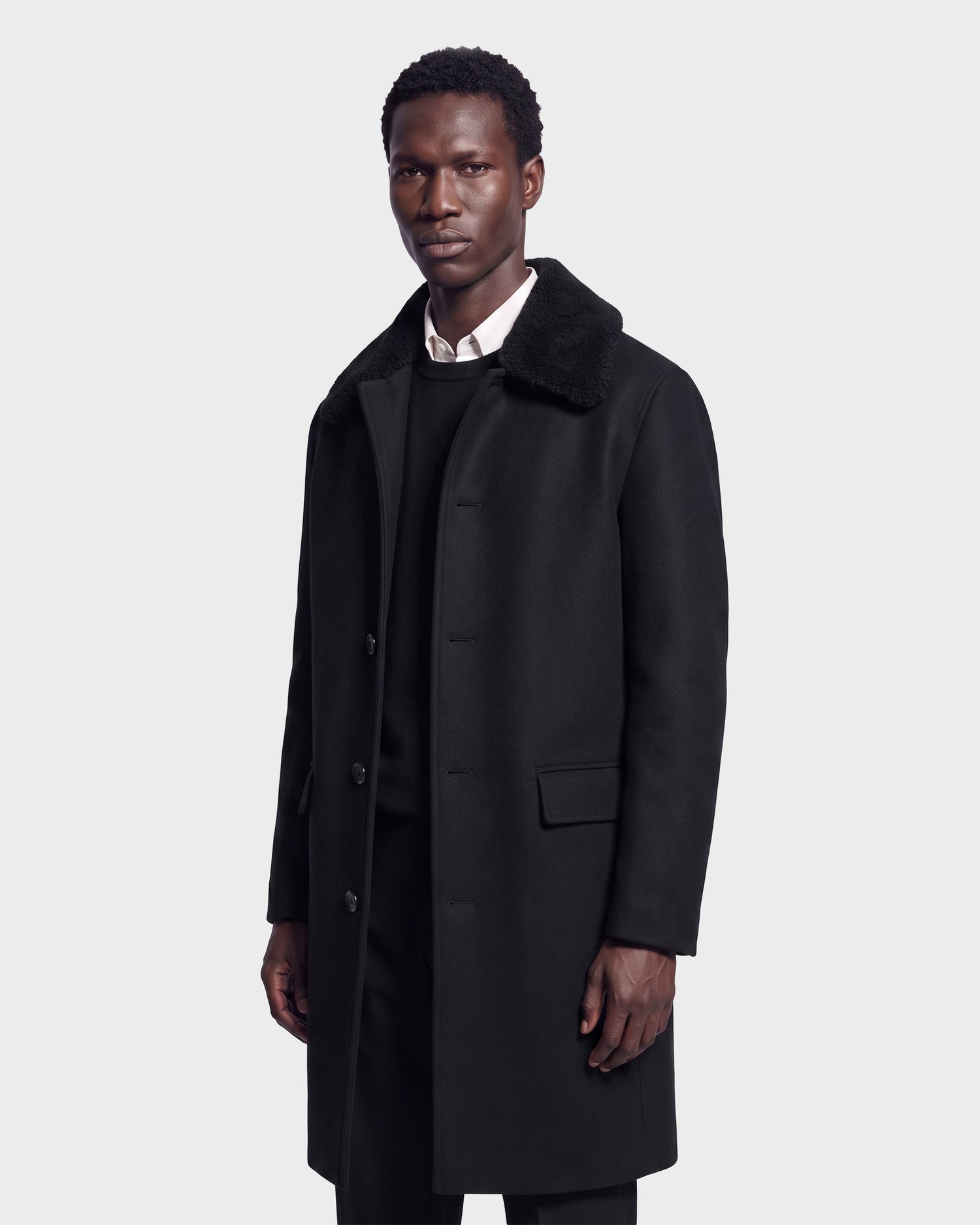 A man wearing the Rudsak NICO - 6126717 Black men’s long wool coat with a removable faux-shearling collar, layered over a black sweater and a white collared shirt, standing against a plain light gray background.