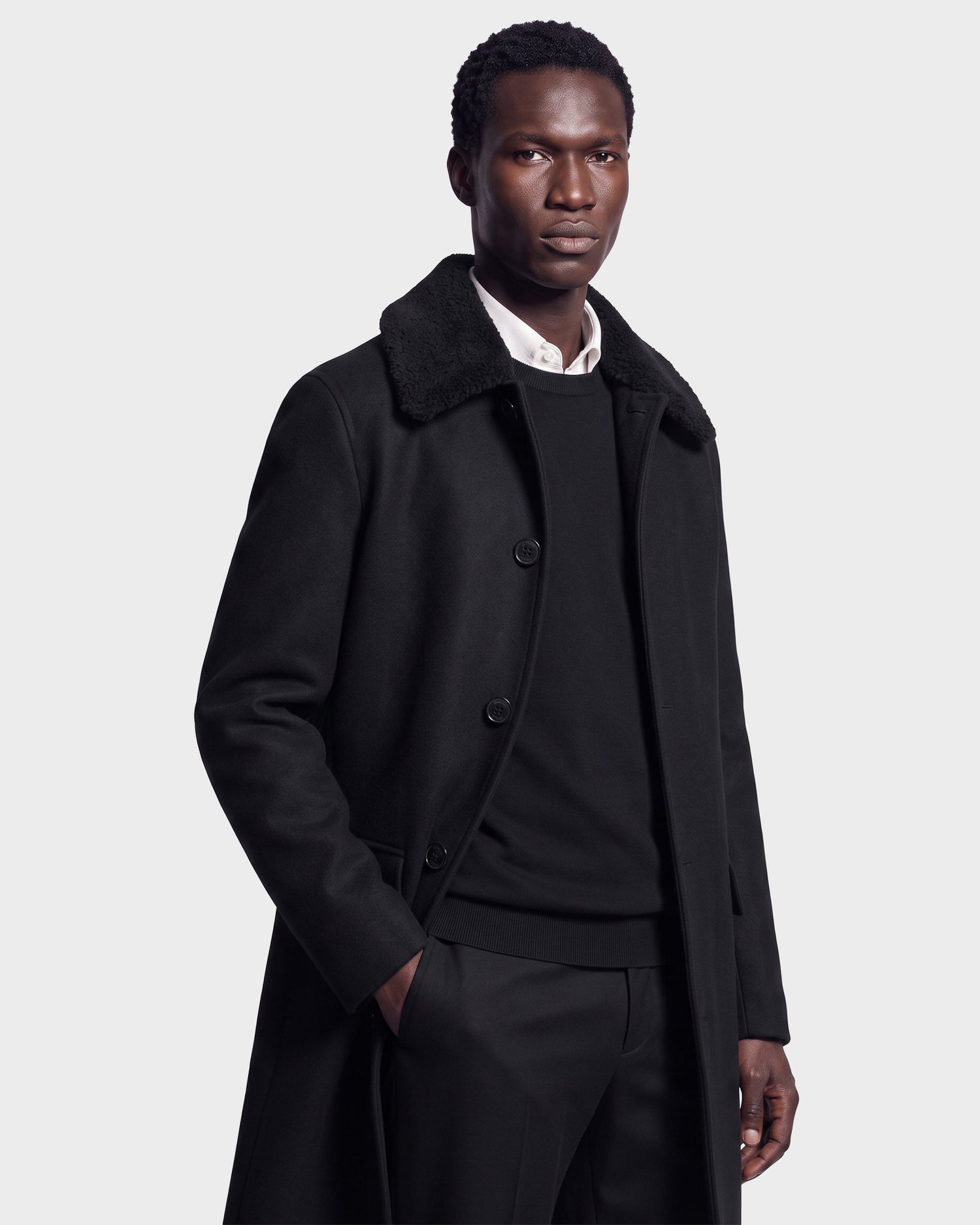 A man stands confidently against a plain light background, wearing the Rudsak NICO - 6126717 Black men’s long wool coat with a removable faux-shearling collar over a black sweater and white shirt, one hand in his pocket as he looks at the camera.