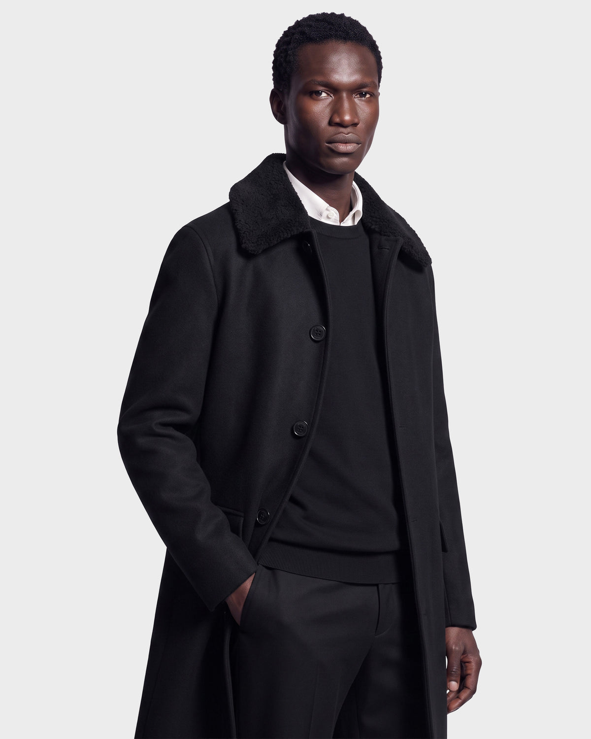 A man stands confidently against a plain light background, wearing the Rudsak NICO - 6126717 Black men’s long wool coat with a removable faux-shearling collar over a black sweater and white shirt, one hand in his pocket as he looks at the camera.