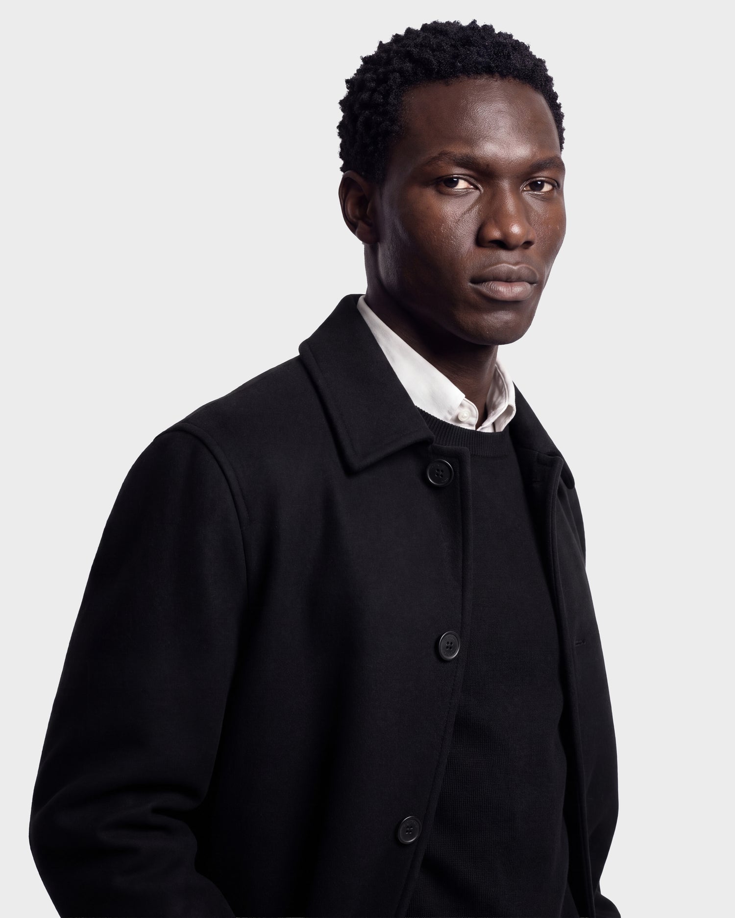 A man with short curly hair wears the Rudsak NICO - 6126717 Black men’s long wool coat with a removable faux-shearling collar, layered over a black sweater and white collared shirt. He stands against a plain light gray background, looking slightly to the side with a serious expression.