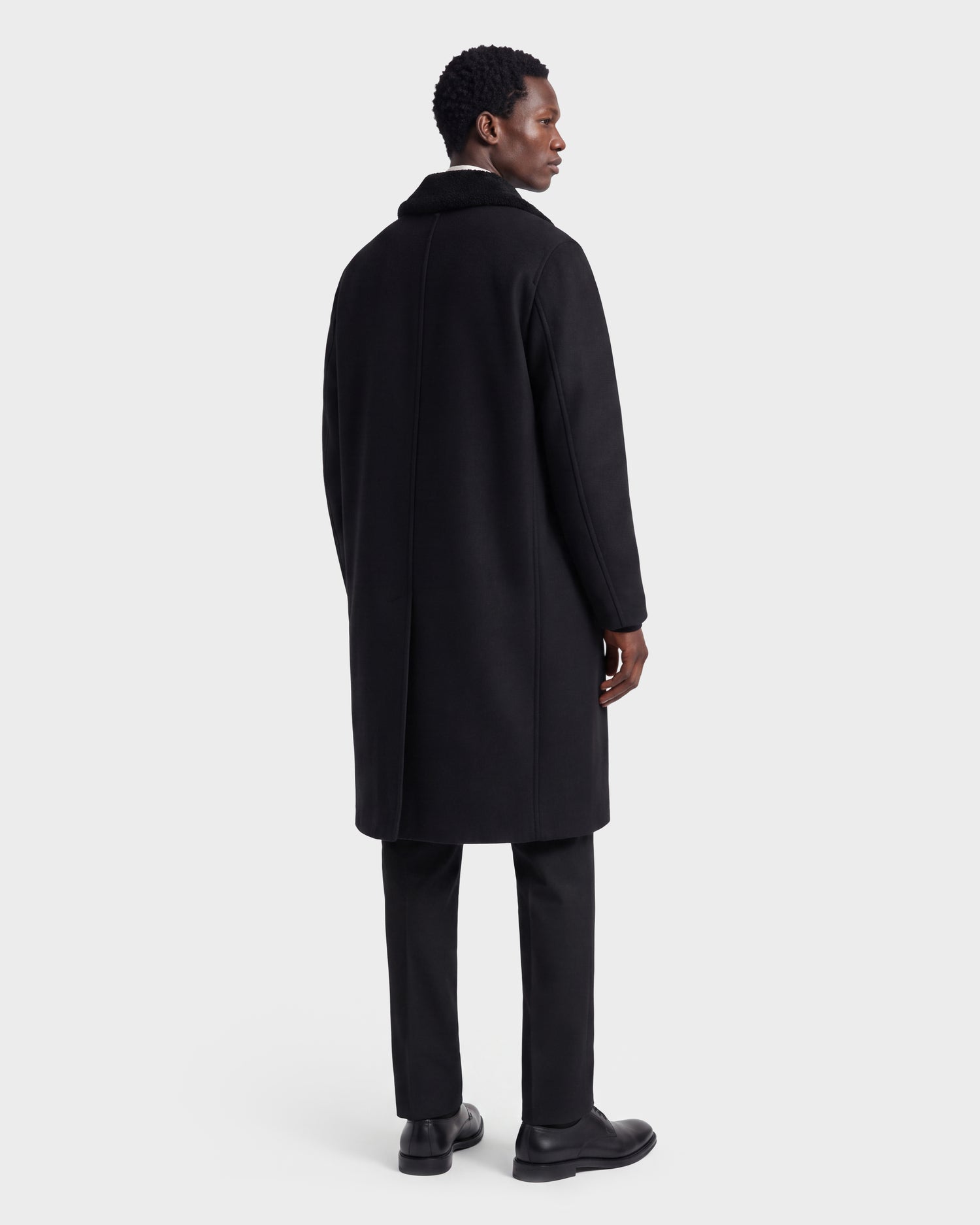 A man stands facing away, wearing the Rudsak NICO - 6126717 Black men’s long wool coat with a removable faux-shearling collar, paired with black pants and black shoes against a plain light gray background.