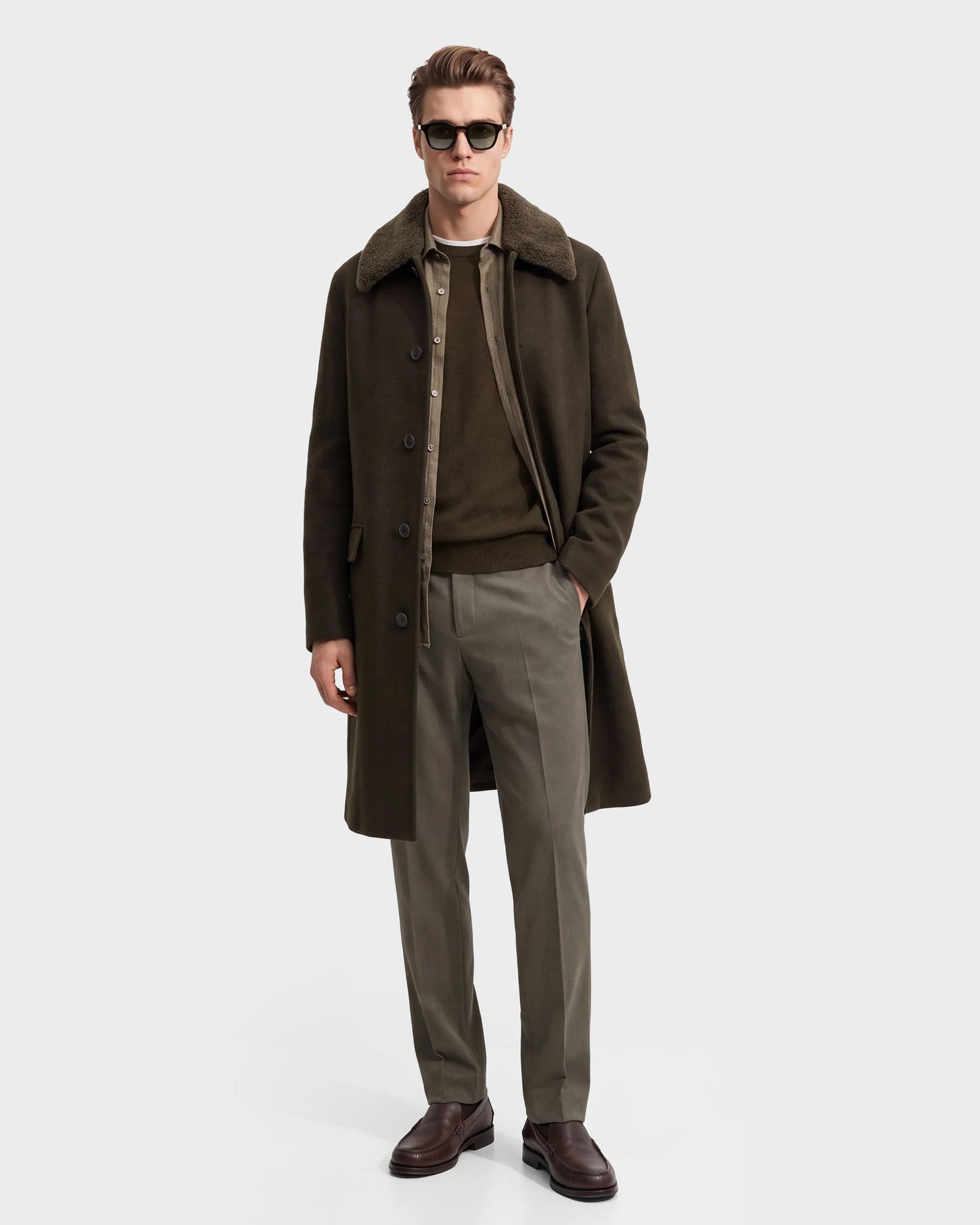 A man wearing sunglasses, Rudsak’s NICO - 6126717 Olive wool blend coat with a shearling collar, an olive sweater, khaki trousers, and brown shoes stands with one hand in his pocket against a plain light background.