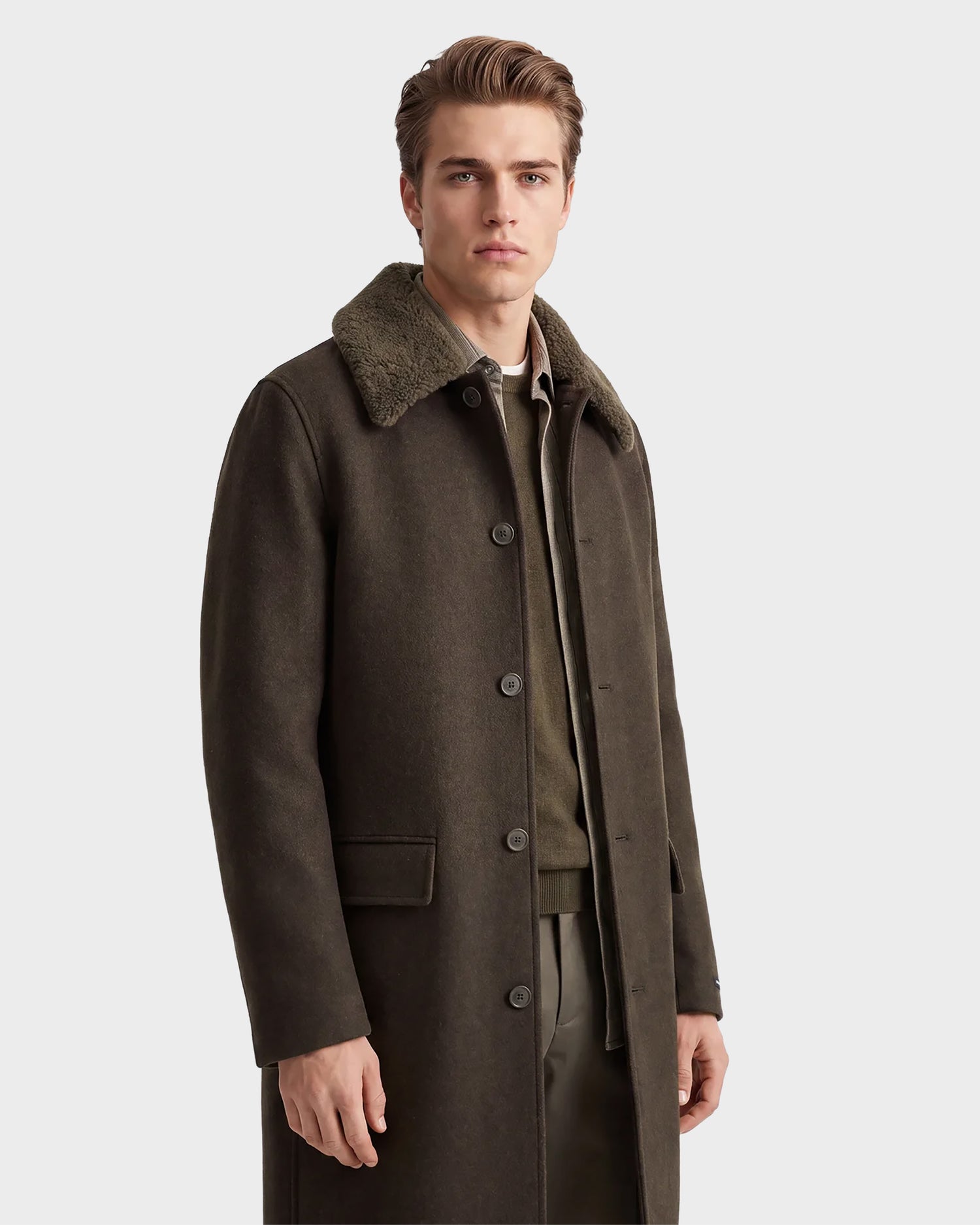 A person stands facing forward against a plain, light background, wearing the Rudsak NICO - 6126717 Olive jacket—a long, dark olive wool-blend coat featuring a fuzzy collar, button closure, and front pockets. The individual has short, styled hair and a neutral expression.