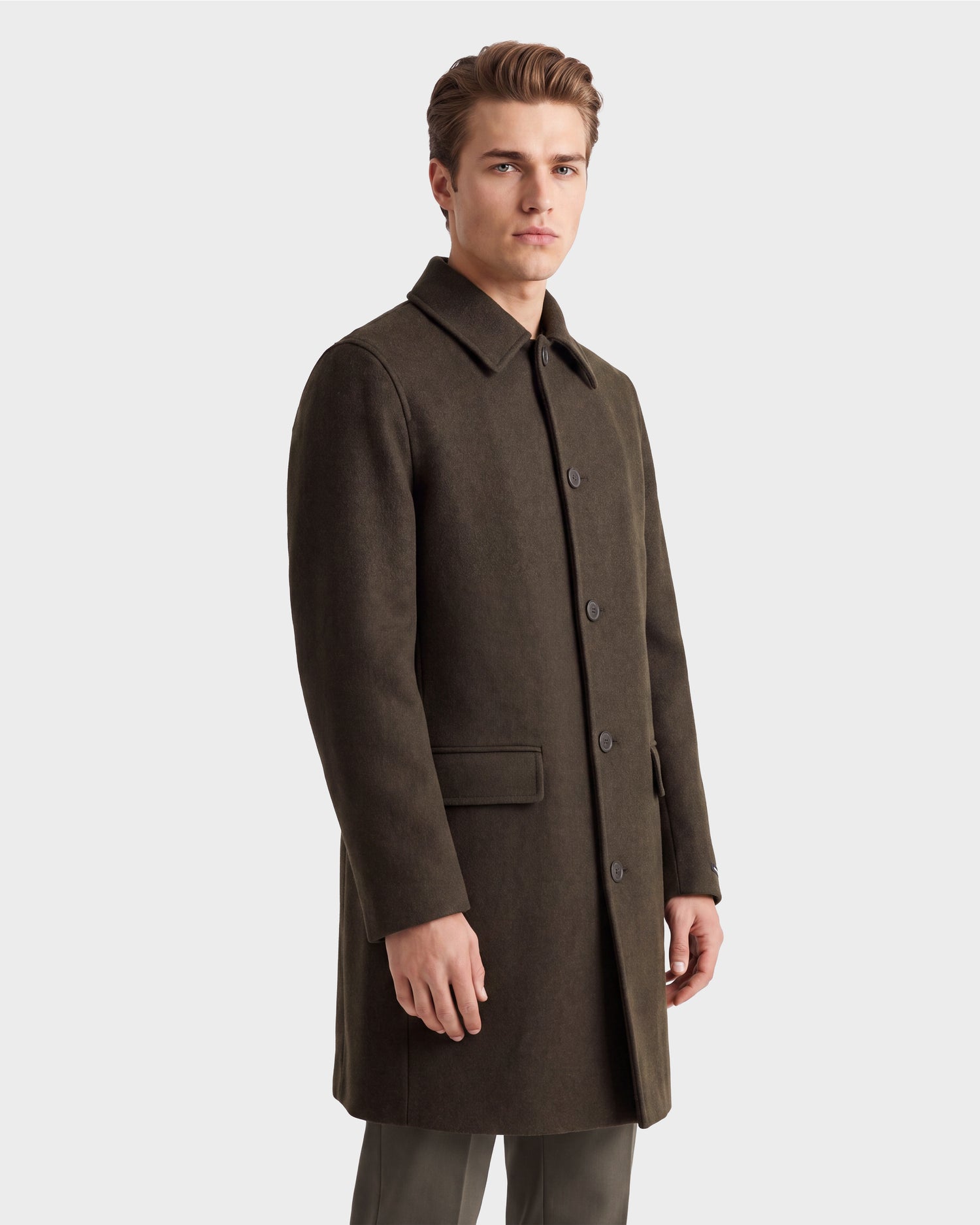 A man wearing the Rudsak NICO - 6126717 Olive jacket, a dark, knee-length wool blend coat with a collar, stands against a plain white background, looking slightly to the side with a neutral expression.