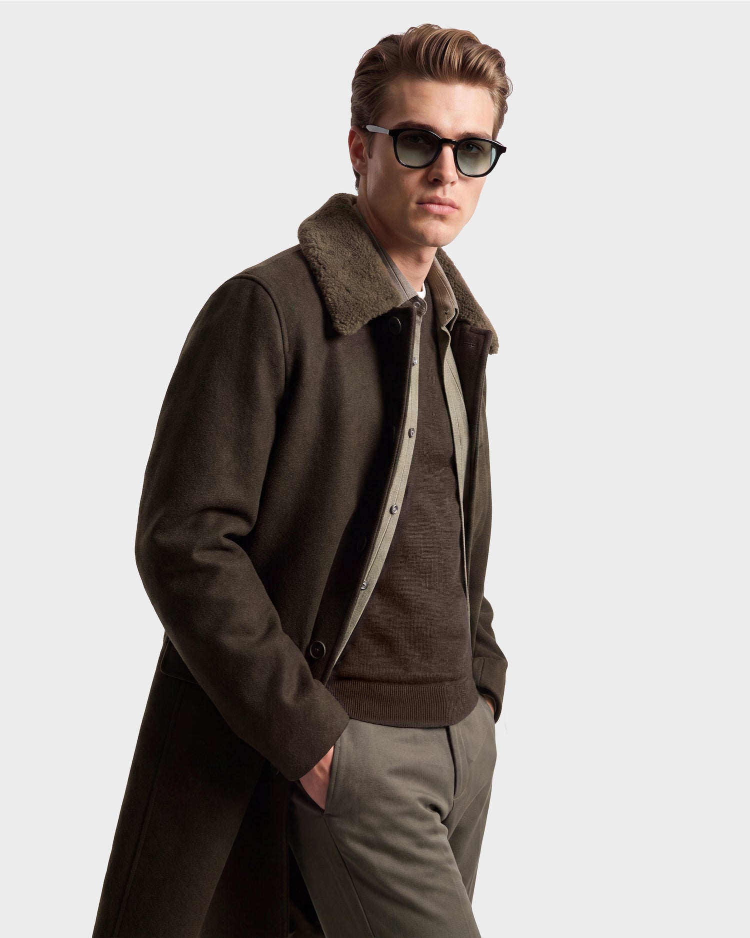 A man wearing sunglasses, an olive green NICO - 6126717 jacket by Rudsak with a fur collar, a brown sweater, and green pants stands with one hand in his pocket against a plain light background.