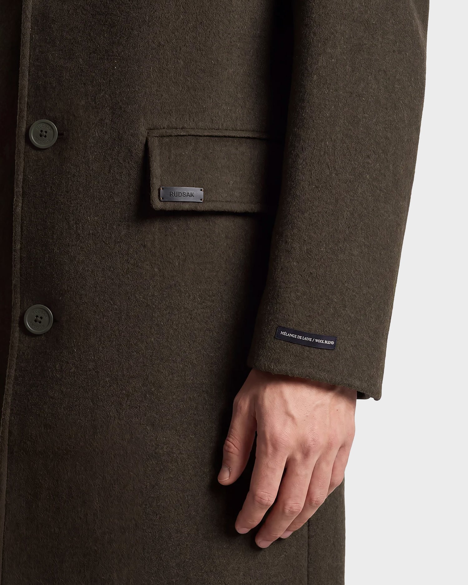 A close-up of a person wearing the Rudsak NICO - 6126717 Olive wool blend coat, featuring buttons, a pocket with a Rudsak label, and their hand near the sleeve adorned with a small tag—perfect for transitional layering.