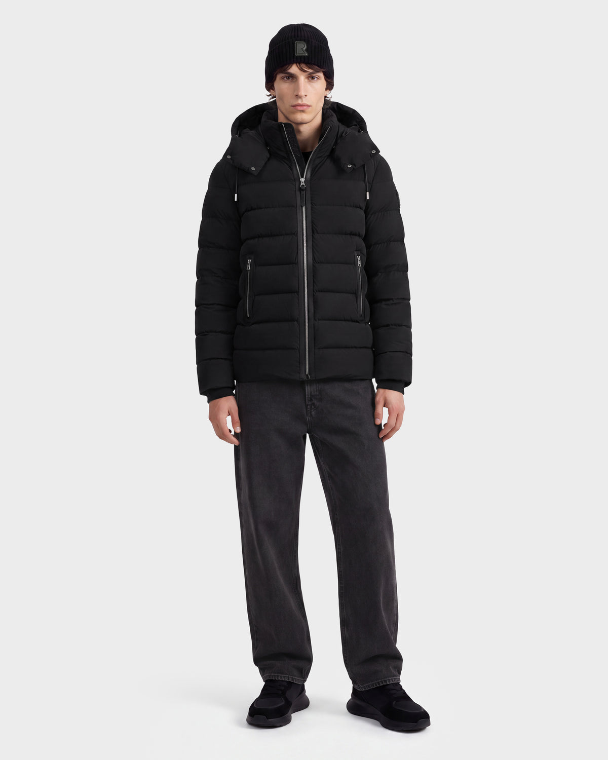 A person wearing the Rudsak NAVAN - 6125598 Black men’s down puffer jacket with RDS-certified down, a black beanie, dark gray jeans, and black shoes stands facing forward against a plain light gray background.