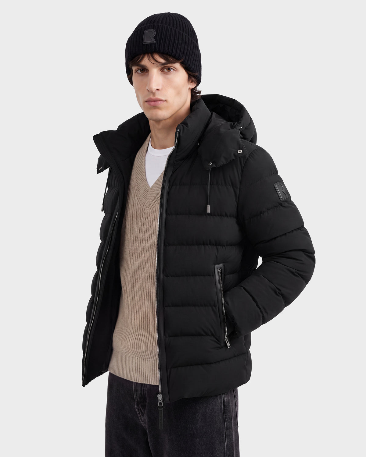 A man wearing the Rudsak NAVAN - 6125598 Black puffer jacket, paired with a beige sweater, white t-shirt, black jeans, and a black beanie poses against a plain light background.