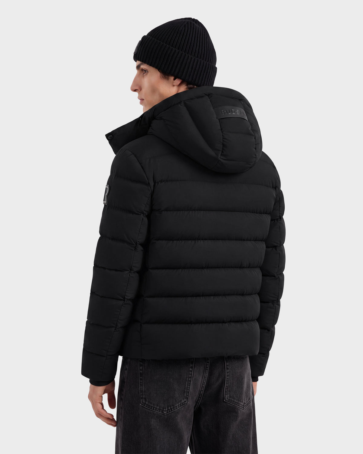 A person wearing the Rudsak NAVAN - 6125598 Black men’s down puffer jacket with a hood, crafted from RDS-certified down, and a black beanie stands facing away against a plain light background.