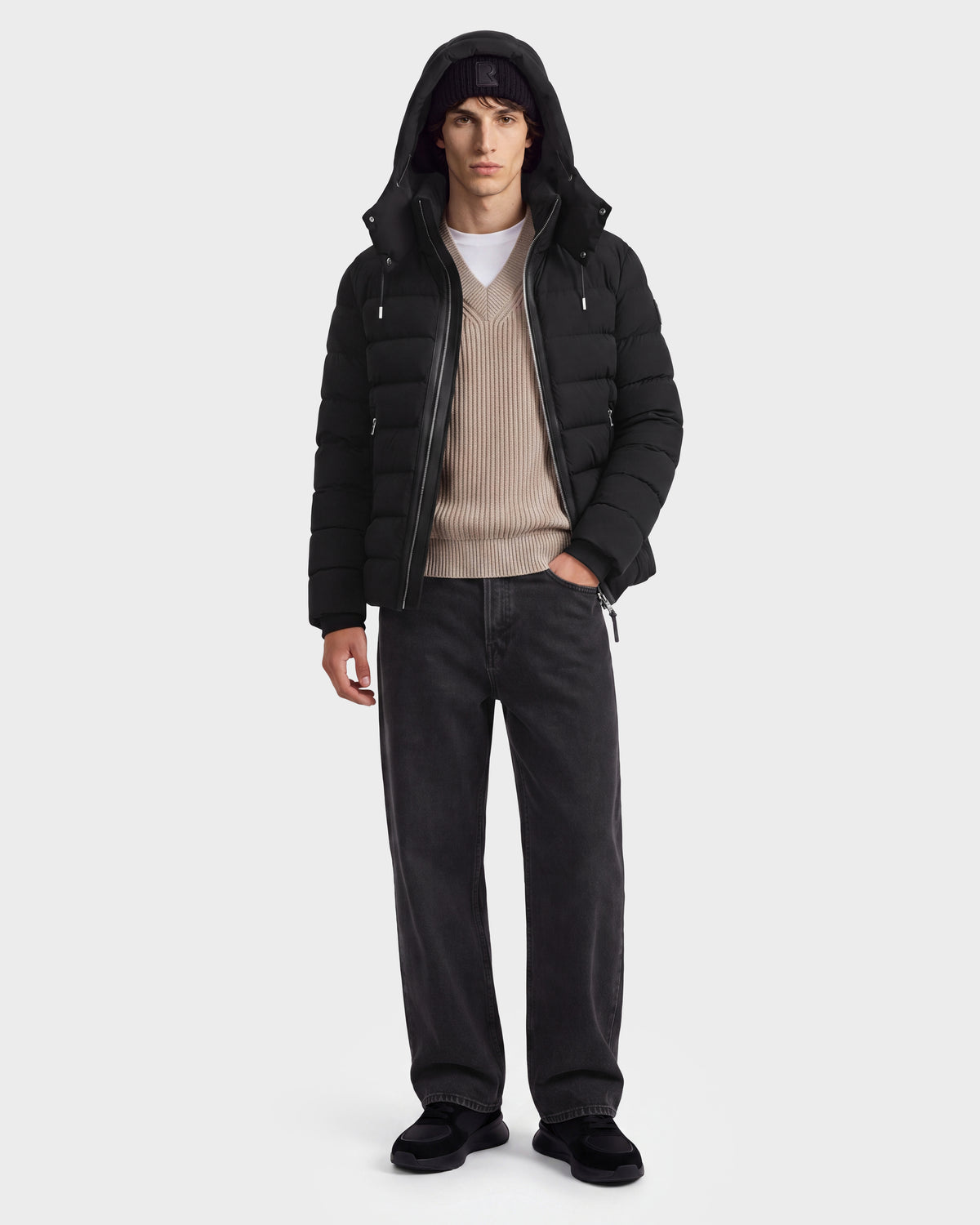 A person wearing the Rudsak NAVAN - 6125598 Black men’s down puffer with RDS-certified down, a beige sweater, white t-shirt, dark jeans, black shoes, and a black beanie stands against a plain white background.