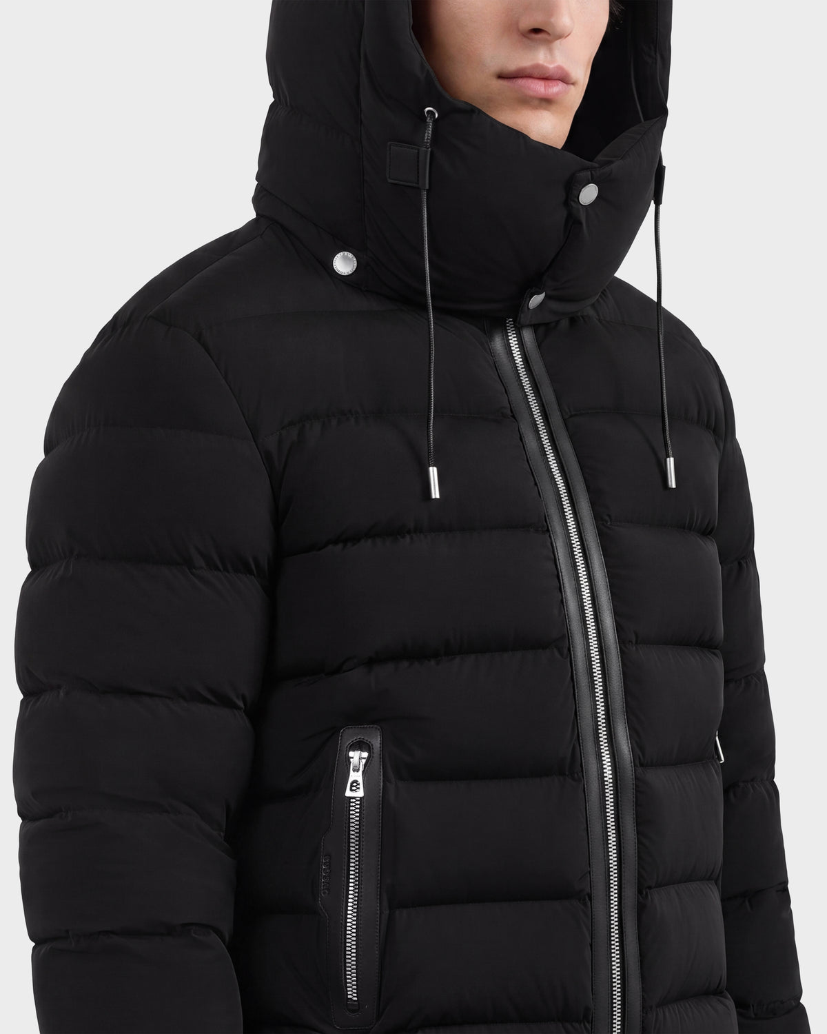 A person shown from the shoulders up against a plain background is wearing the Rudsak NAVAN - 6125598 Black men’s down puffer jacket, featuring a hood, silver zippers, and metal snap buttons.