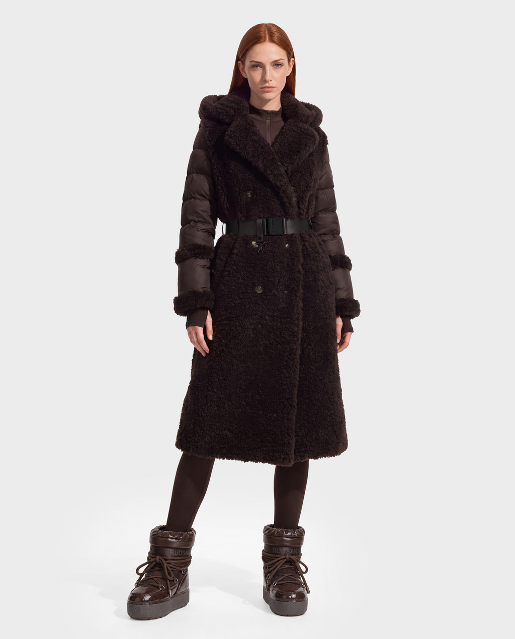 A model wears the NOELLE - 8126971 Espresso, a knee-length, dark brown hooded coat featuring a combination of quilted puffer sleeves and a fuzzy faux fur body with a buckled belt.