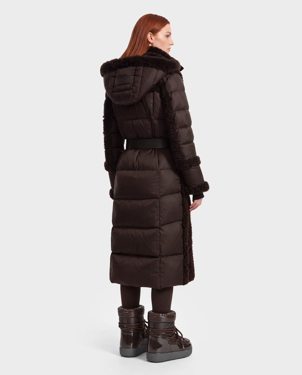 A person seen from the back wearing a dark brown, belted, faux fur hooded NOELLE - 8126971 Espresso down coat, paired with matching brown boots.