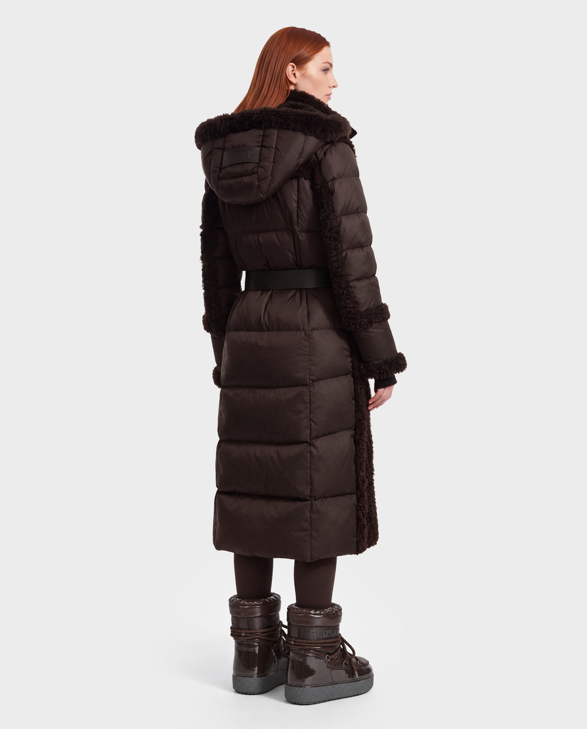 A person seen from the back wearing a dark brown, belted, faux fur hooded NOELLE - 8126971 Espresso down coat, paired with matching brown boots.