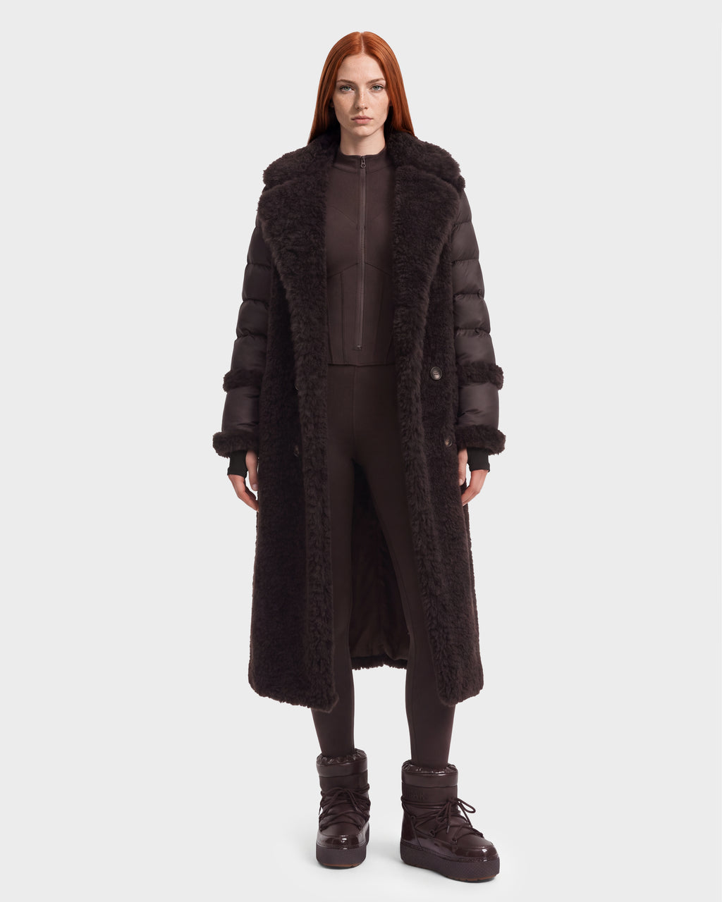A woman wearing a NOELLE - 8126971 Espresso hooded, dark brown down puffer coat with shearling trim over a matching zip-up jumpsuit and dark brown winter boots.