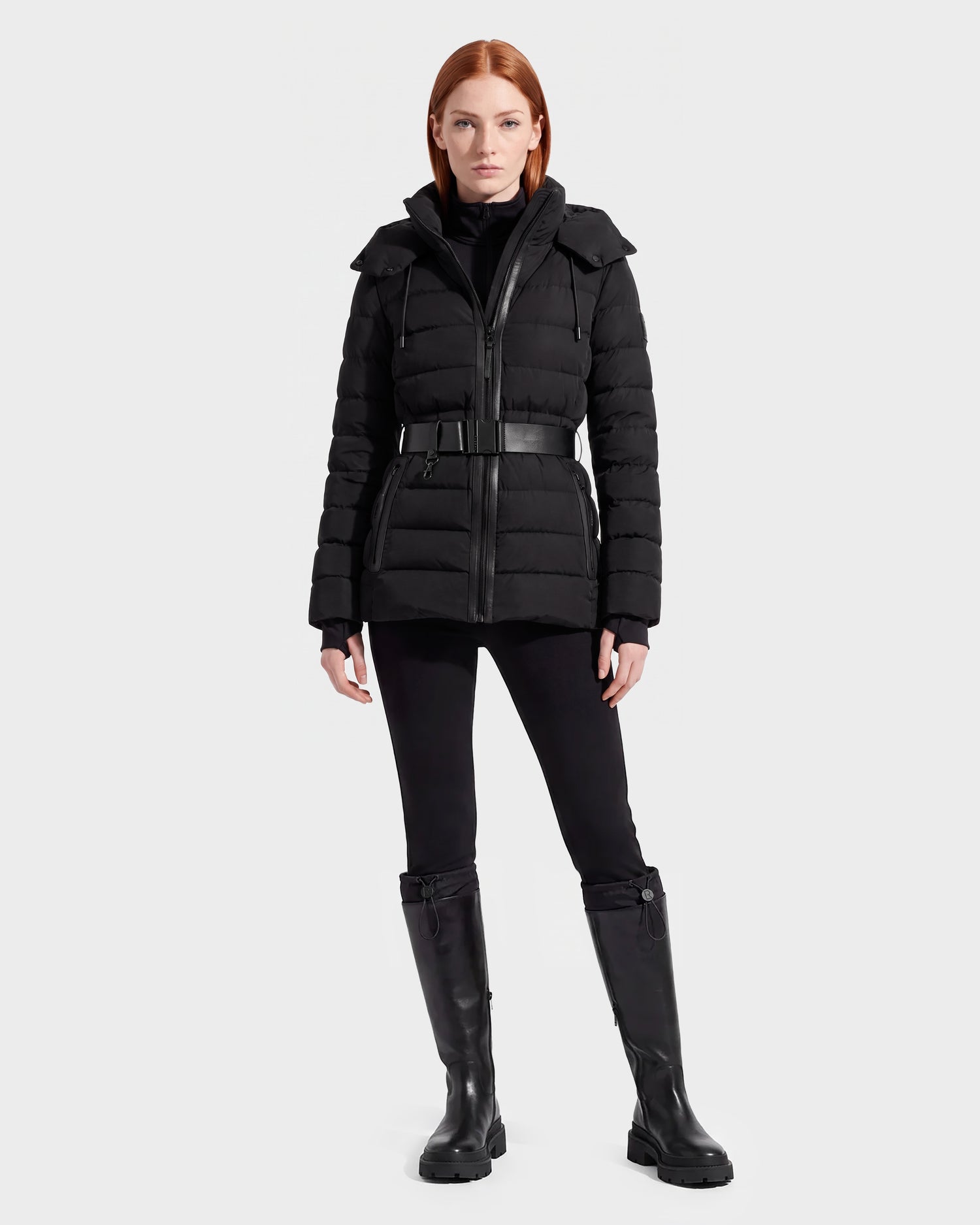 A woman with straight red hair wears the Rudsak PELIN S - 8124813 Black belted quilted puffer jacket, paired with black pants and knee-high black boots, standing against a plain light gray background.