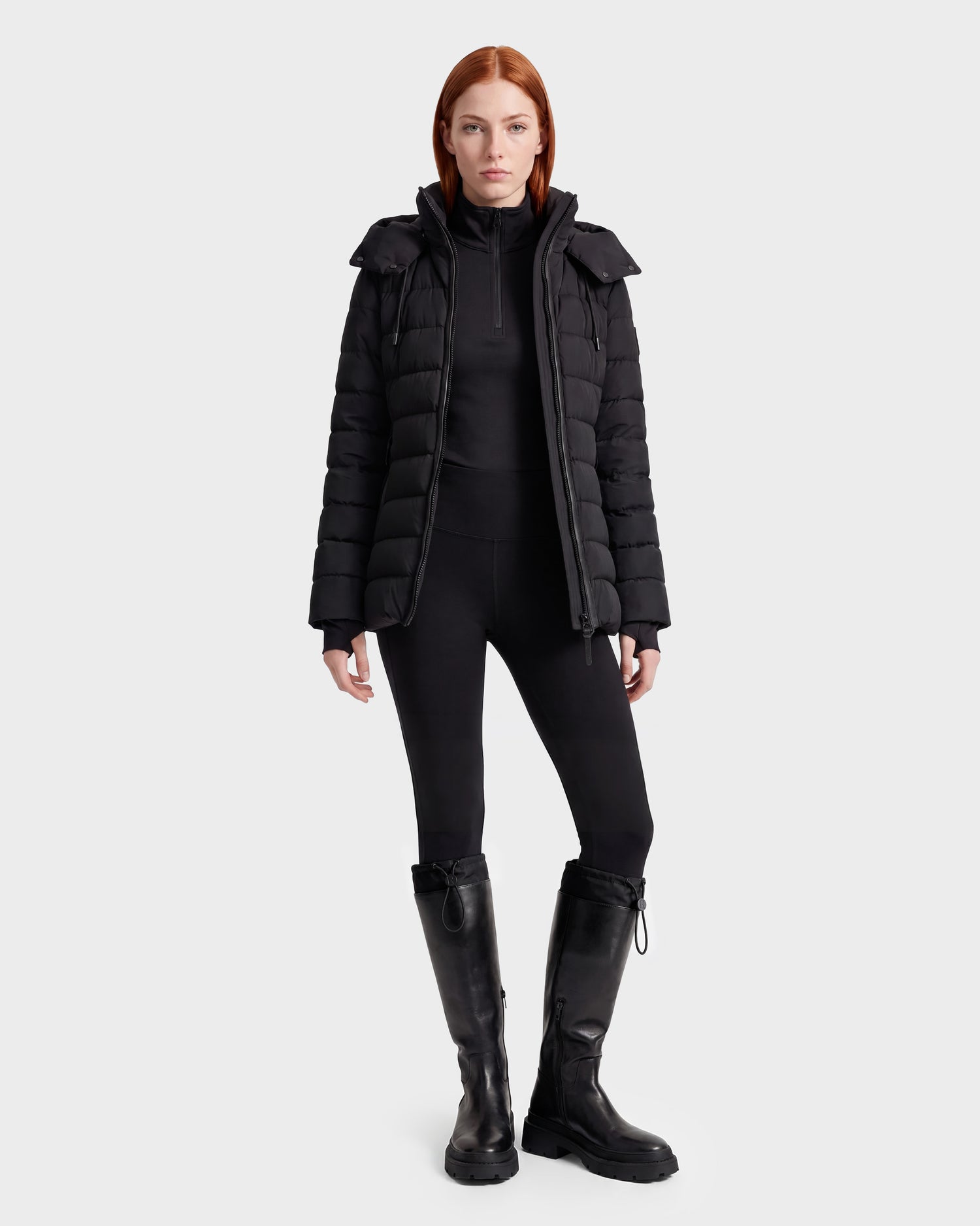 A woman with straight red hair wears the Rudsak PELIN S - 8124813 Black quilted puffer jacket, paired with black leggings, a black top, and black knee-high boots, standing against a plain light background.