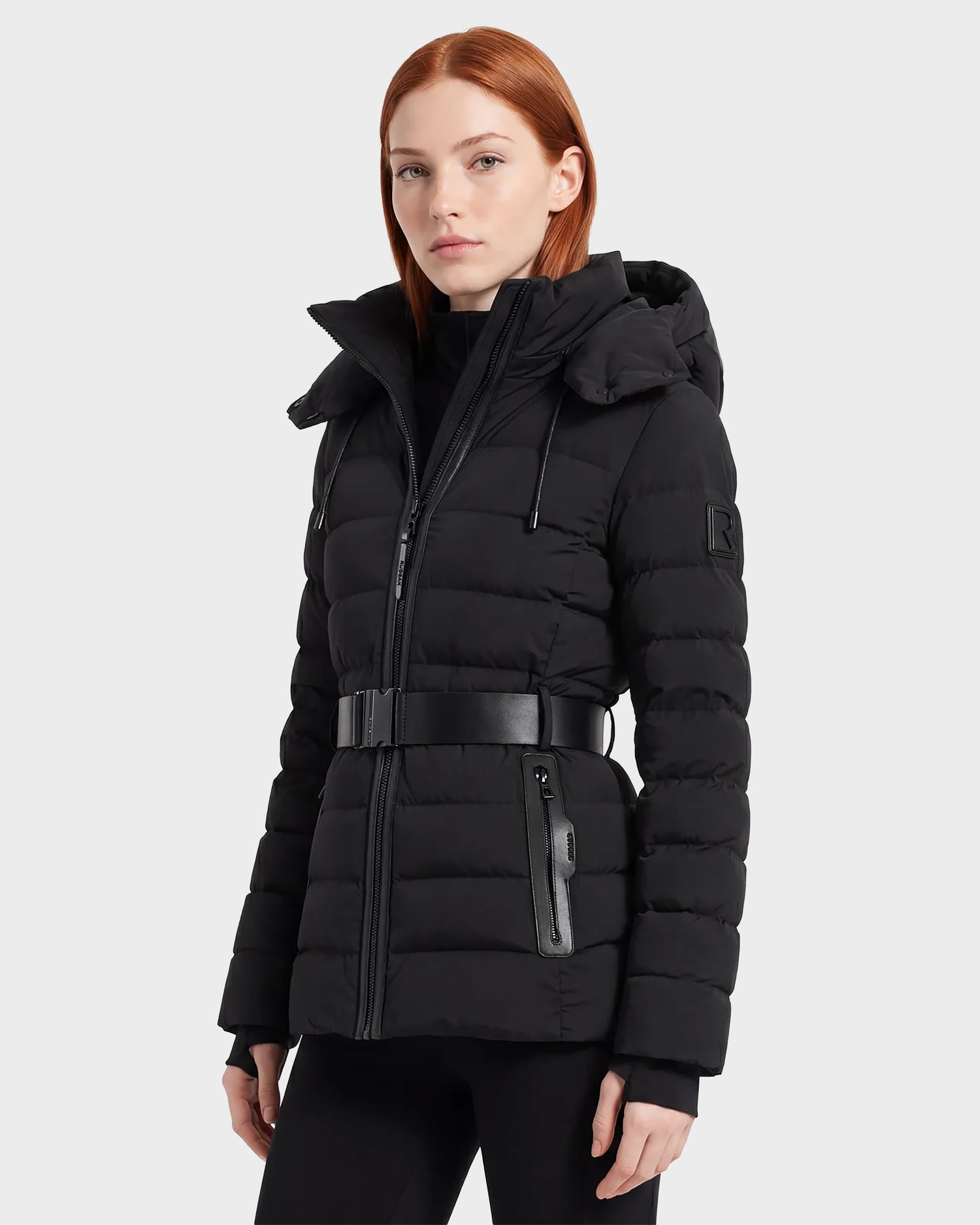 A woman with straight red hair is wearing the Rudsak PELIN S - 8124813 Black, a belted black quilted puffer jacket with a hood, standing against a plain light gray background.