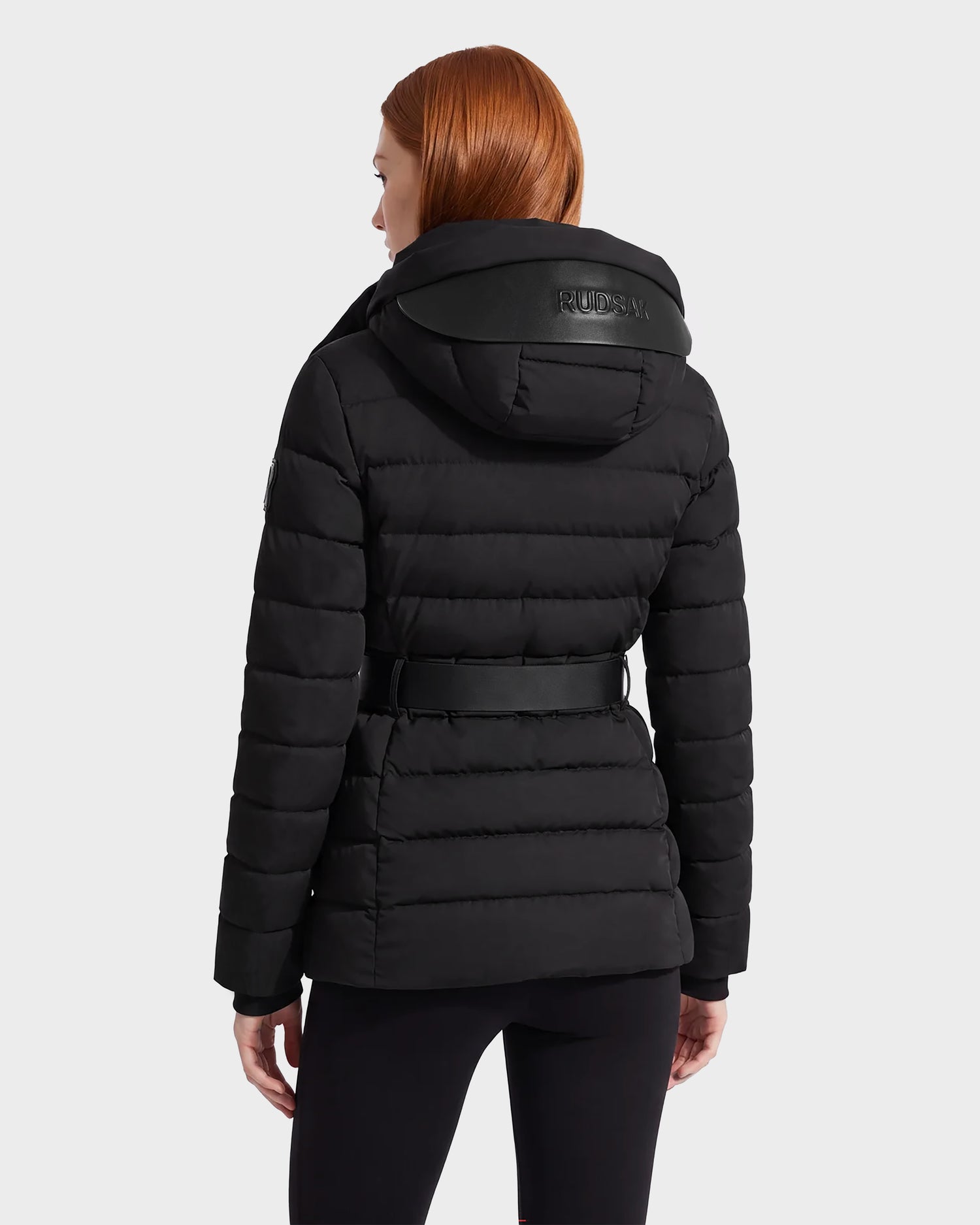 A person with straight red hair is seen from the back against a plain gray background, wearing the Rudsak PELIN S - 8124813 Black women's down puffer jacket with a belt and hood.