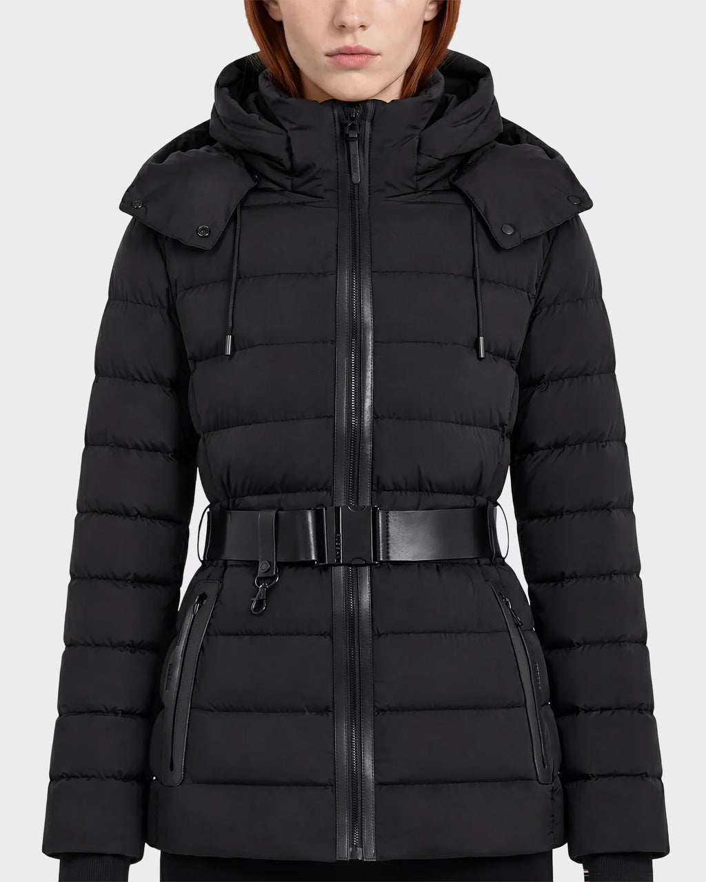 A woman wearing the Rudsak PELIN S - 8124813 Black coat, a belted quilted puffer jacket with a hood, zippered front, and zippered pockets, stands against a plain light background.