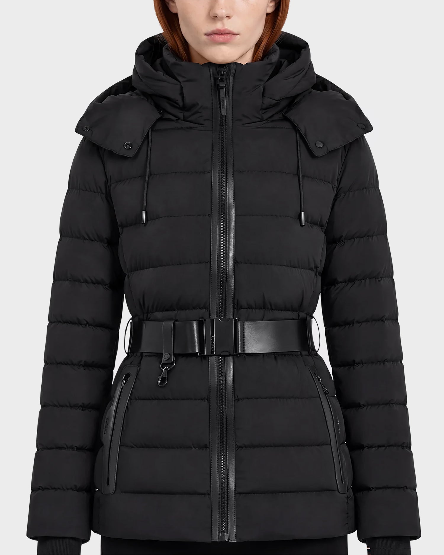 A woman wearing the Rudsak PELIN S - 8124813 Black coat, a belted quilted puffer jacket with a hood, zippered front, and zippered pockets, stands against a plain light background.