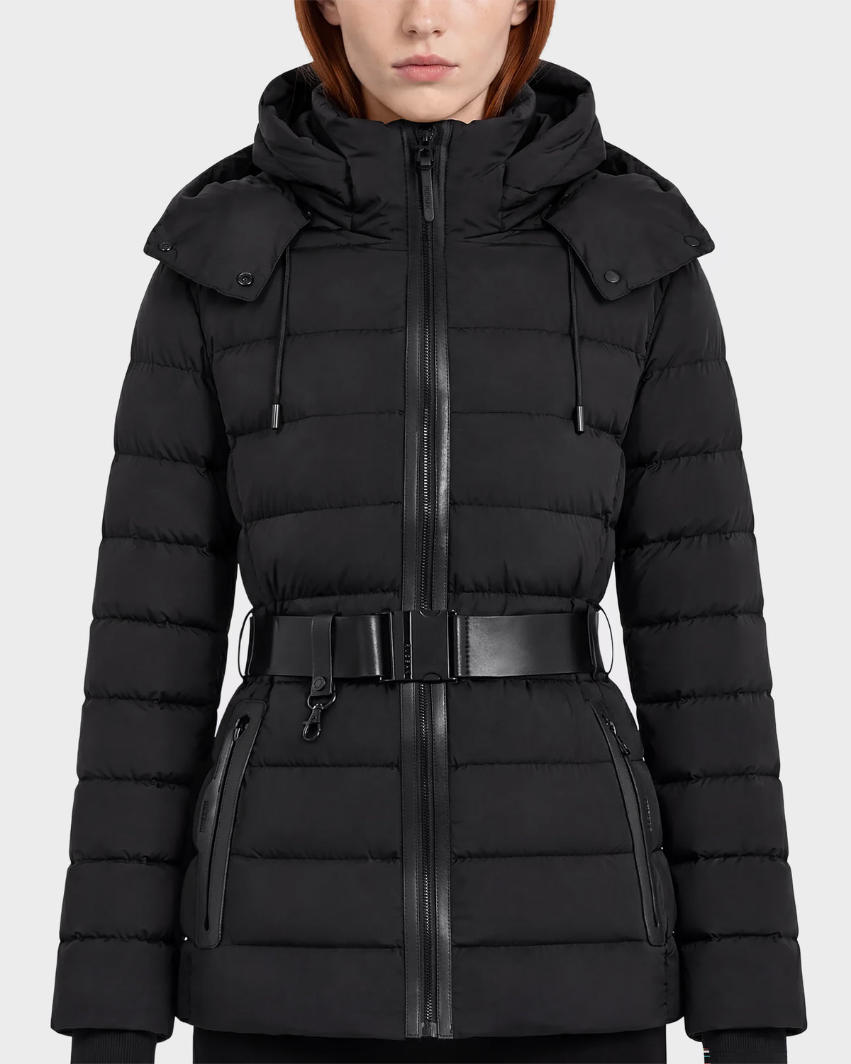 A woman wearing the Rudsak PELIN S - 8124813 Black coat, a belted quilted puffer jacket with a hood, zippered front, and zippered pockets, stands against a plain light background.