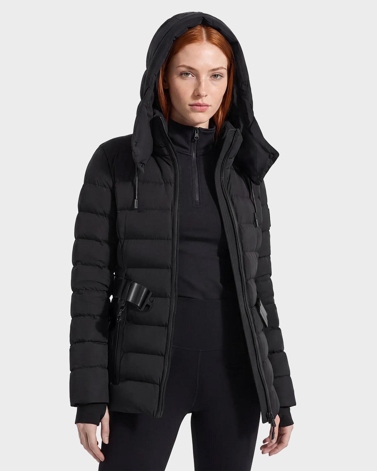 A woman with straight red hair wears the Rudsak PELIN S - 8124813 Black quilted puffer jacket, styled with a black zip-up top and black pants, standing against a plain light gray background.