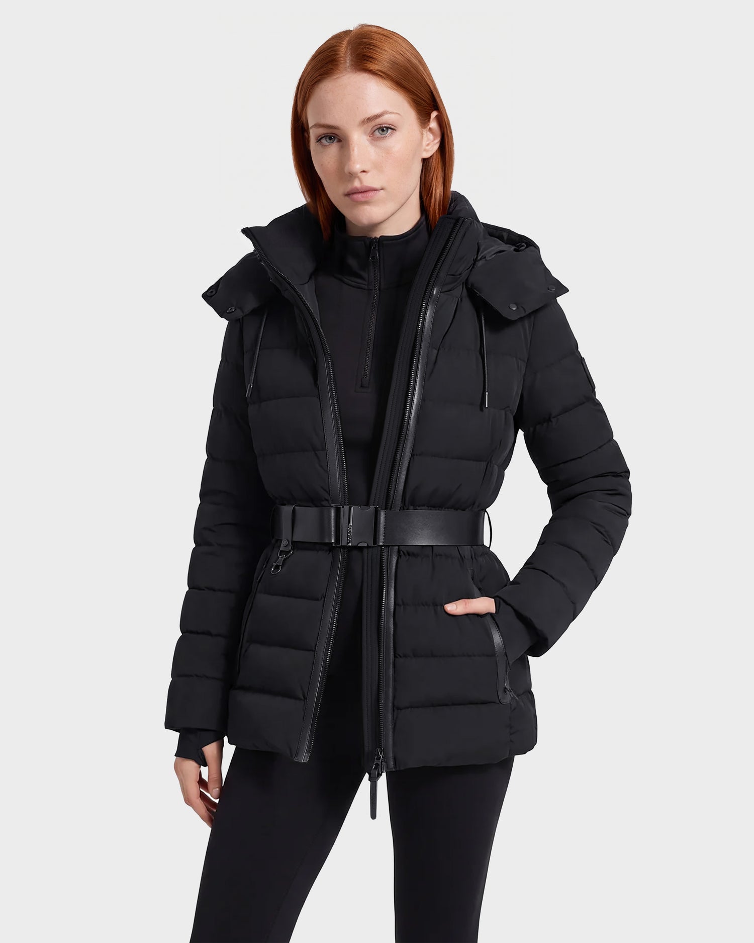 A woman with straight red hair wears the Rudsak PELIN S - 8124813 black belted quilted puffer jacket over a black top and black pants, standing against a plain, light background.