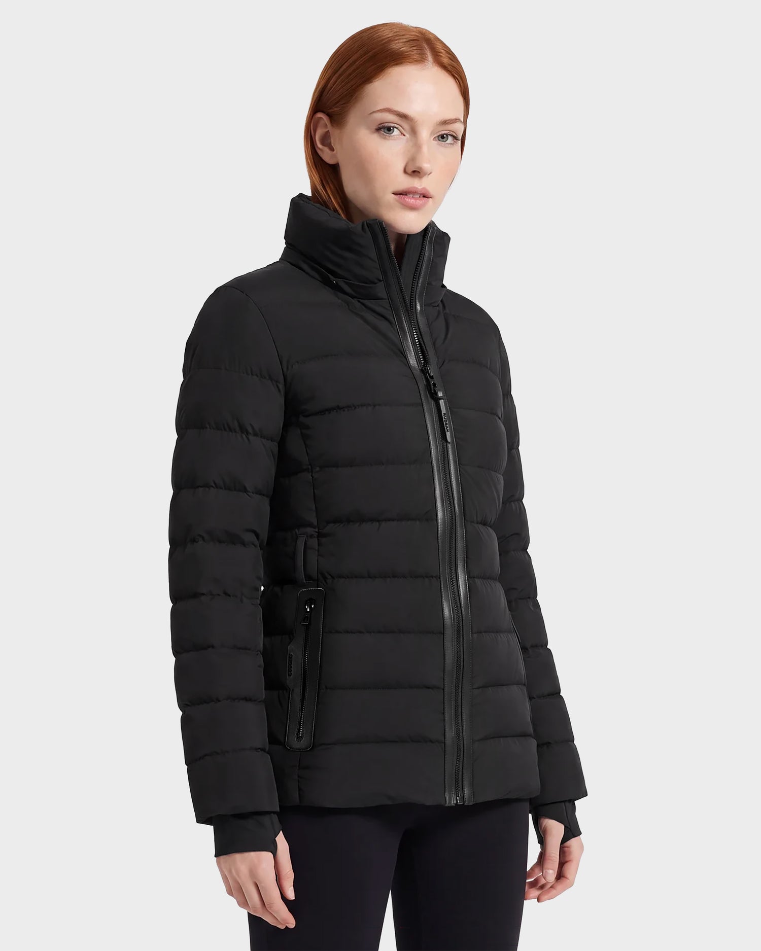 A woman with straight red hair models the Rudsak PELIN S - 8124813 Black women's down puffer jacket, featuring a high collar and zippered pockets, standing against a plain light background.