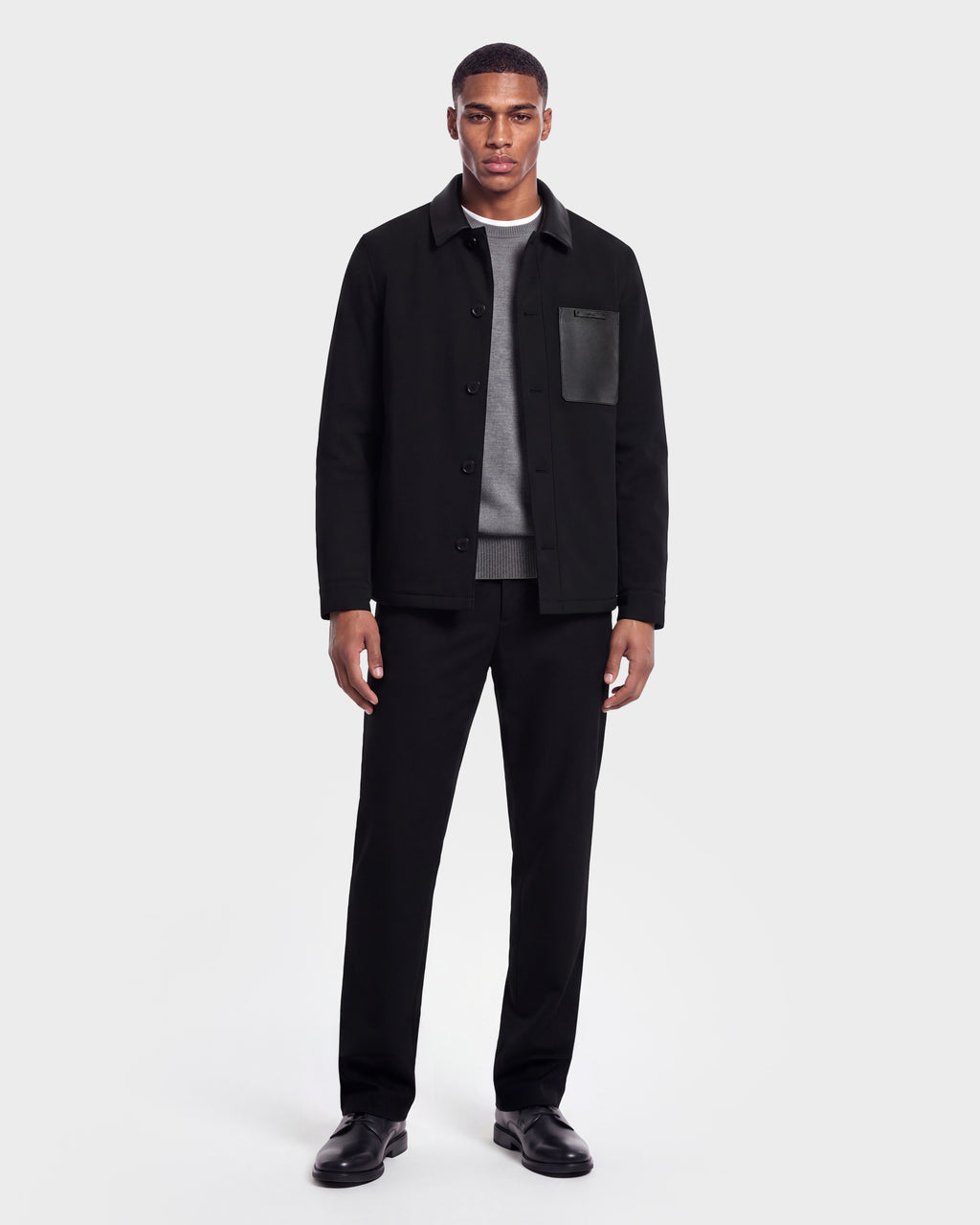 A man stands facing forward wearing the Rudsak PALMER - 6125002 Black shirt jacket featuring eco-fill insulation and subtle leather detailing, paired with a gray sweater, black pants, and black shoes against a plain white background.