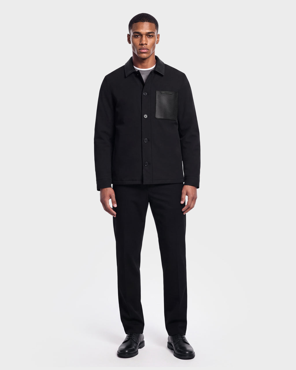 A man stands against a white background wearing the Rudsak PALMER - 6125002 Black modern shirt jacket with subtle leather detailing and a chest pocket, paired with black pants, a gray shirt, and black shoes.