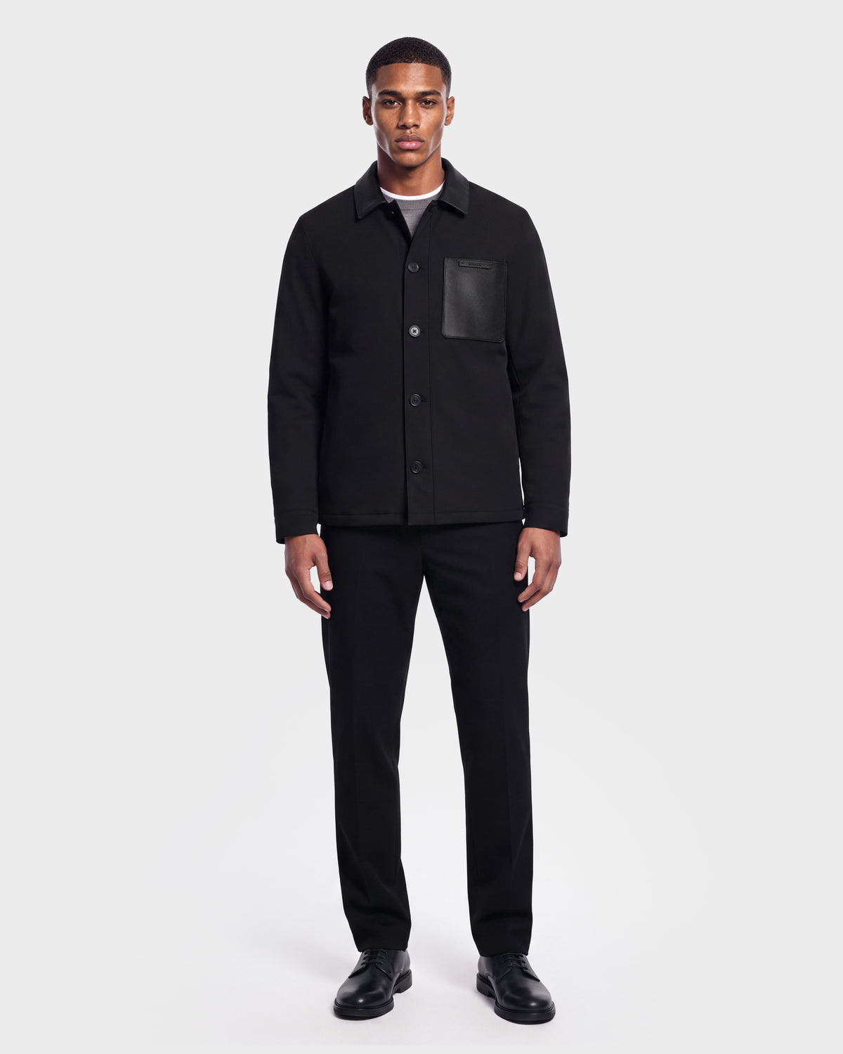A man stands against a white background wearing the Rudsak PALMER - 6125002 Black modern shirt jacket with subtle leather detailing and a chest pocket, paired with black pants, a gray shirt, and black shoes.