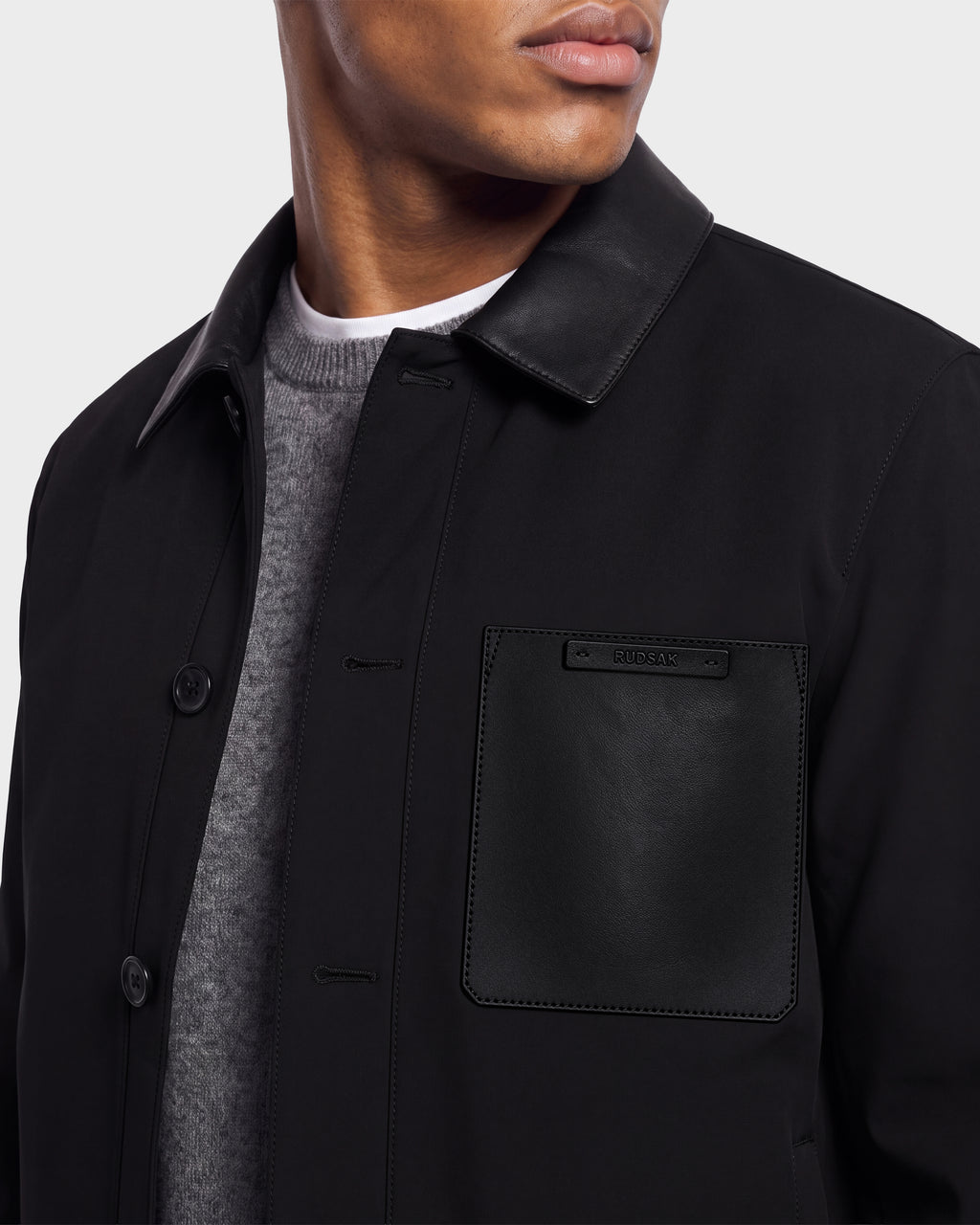 A person is shown from the shoulders to mid-chest wearing the Rudsak PALMER - 6125002 Black—a modern men’s shirt jacket featuring a leather collar and leather chest pocket—layered over a gray sweater.