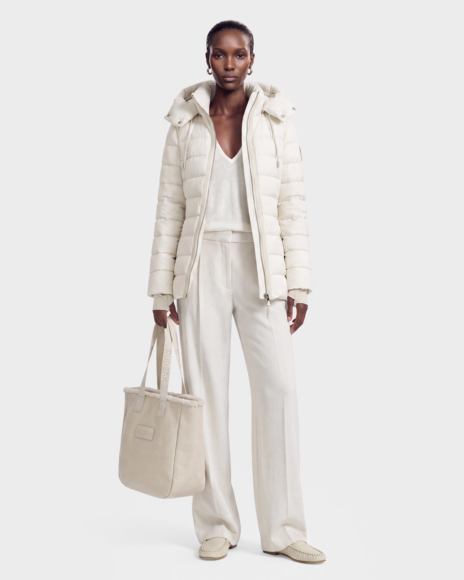 A woman stands against a white background, holding the Rudsak PELIN S - 8124813 Vanilla quilted jacket and matching cream tote bag, styled with a V-neck top, wide-leg pants, and loafers.