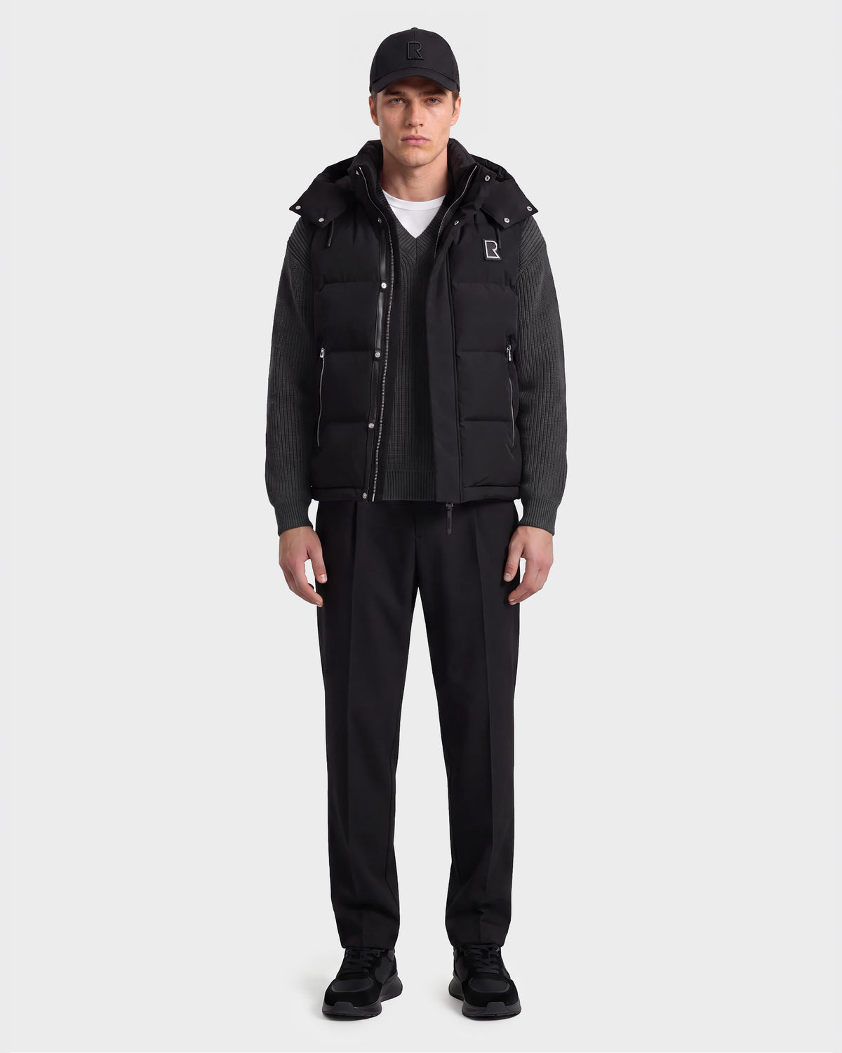 A man stands facing forward wearing the black Rudsak REN M - 6125691 men’s down puffer vest layered over a gray sweater, paired with a black cap, black pants, and black shoes against a plain white background.
