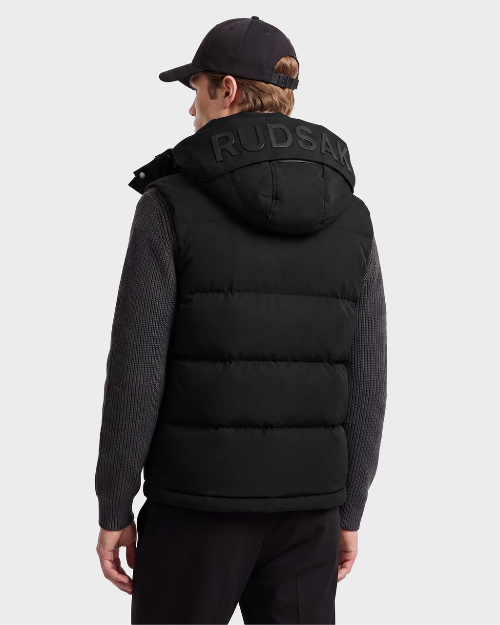 A person wearing the Rudsak REN M - 6125691 Black men’s down puffer vest, featuring RUDSAK branding on the hood, pairs it with a dark sweater, black pants, and a black cap while facing away from the camera against a plain background.