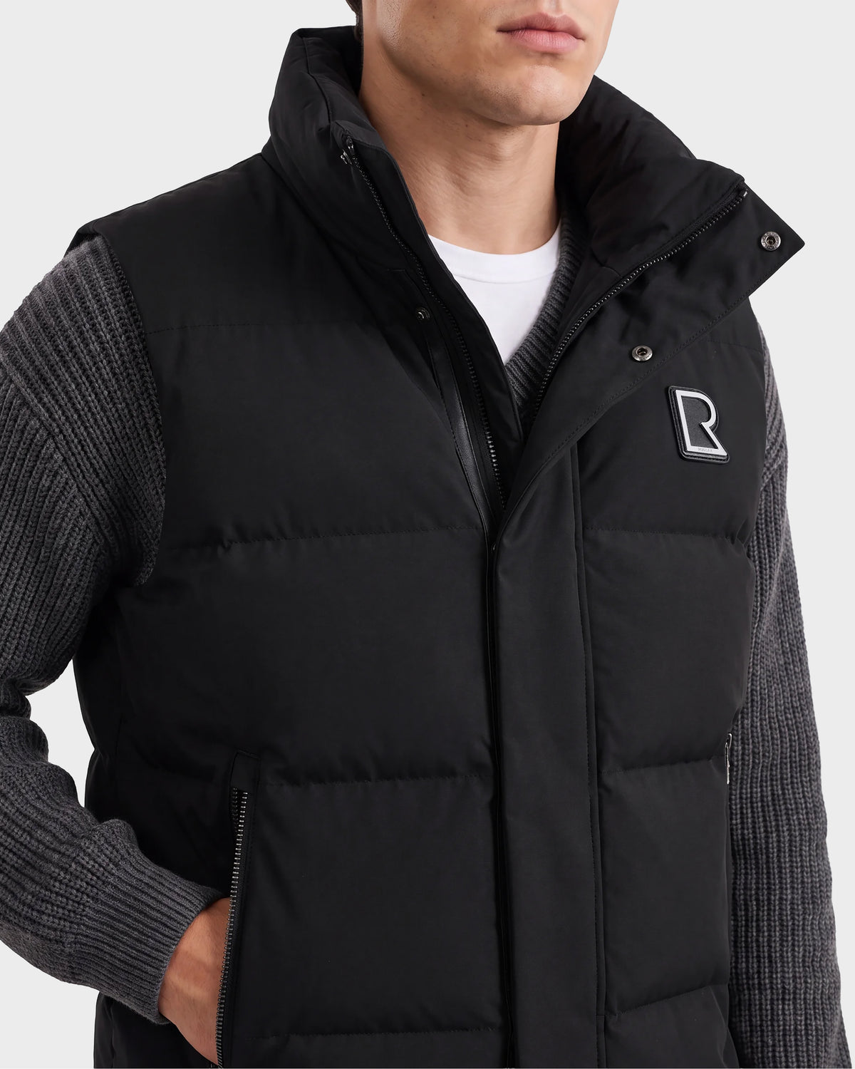 A man is shown from the shoulders to the waist, wearing a black Rudsak REN M - 6125691 down puffer vest over a gray ribbed sweater. The quilted vest features an R logo on the left chest and is zipped up.
