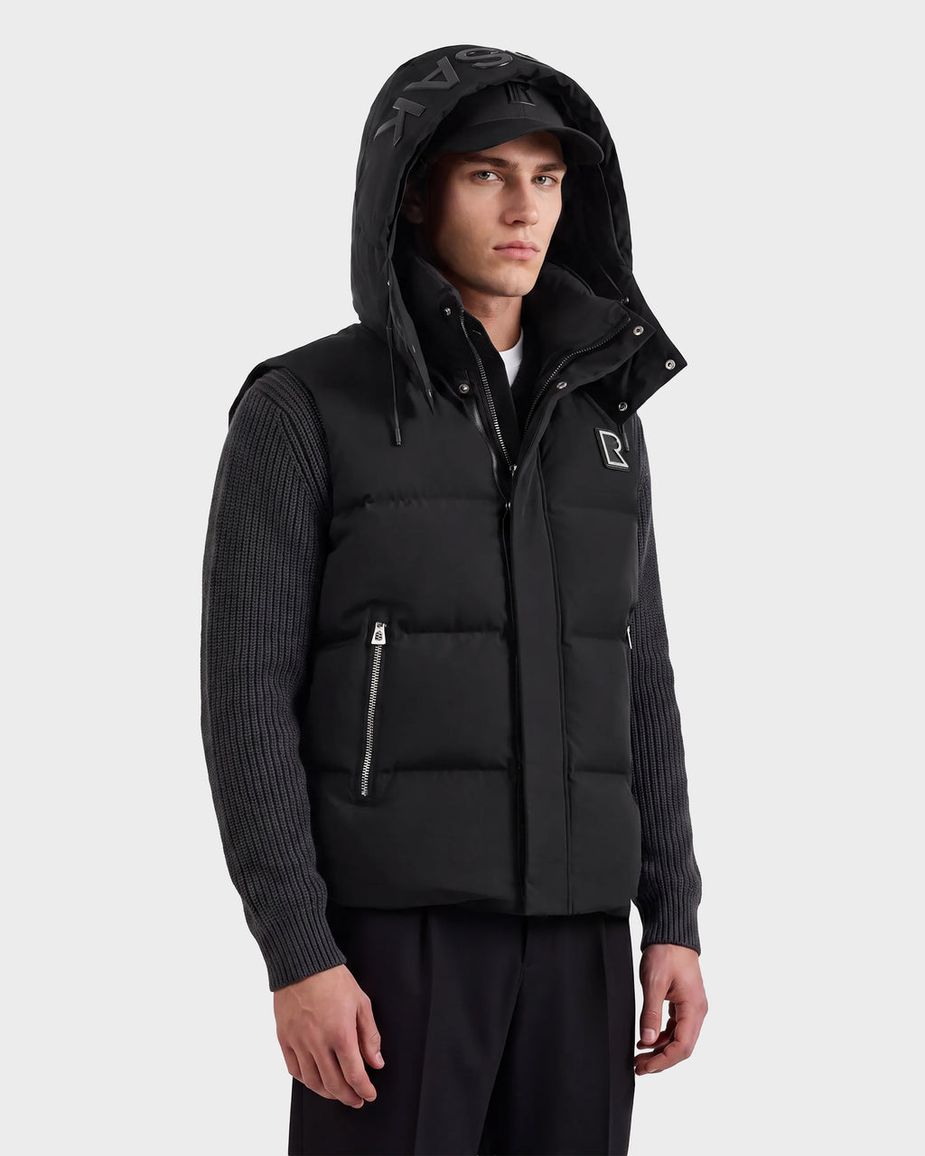 A man wearing the Rudsak REN M - 6125691 Black quilted vest with a hood over a ribbed dark sweater and black pants stands sideways, looking at the camera with a neutral expression against a plain light background.