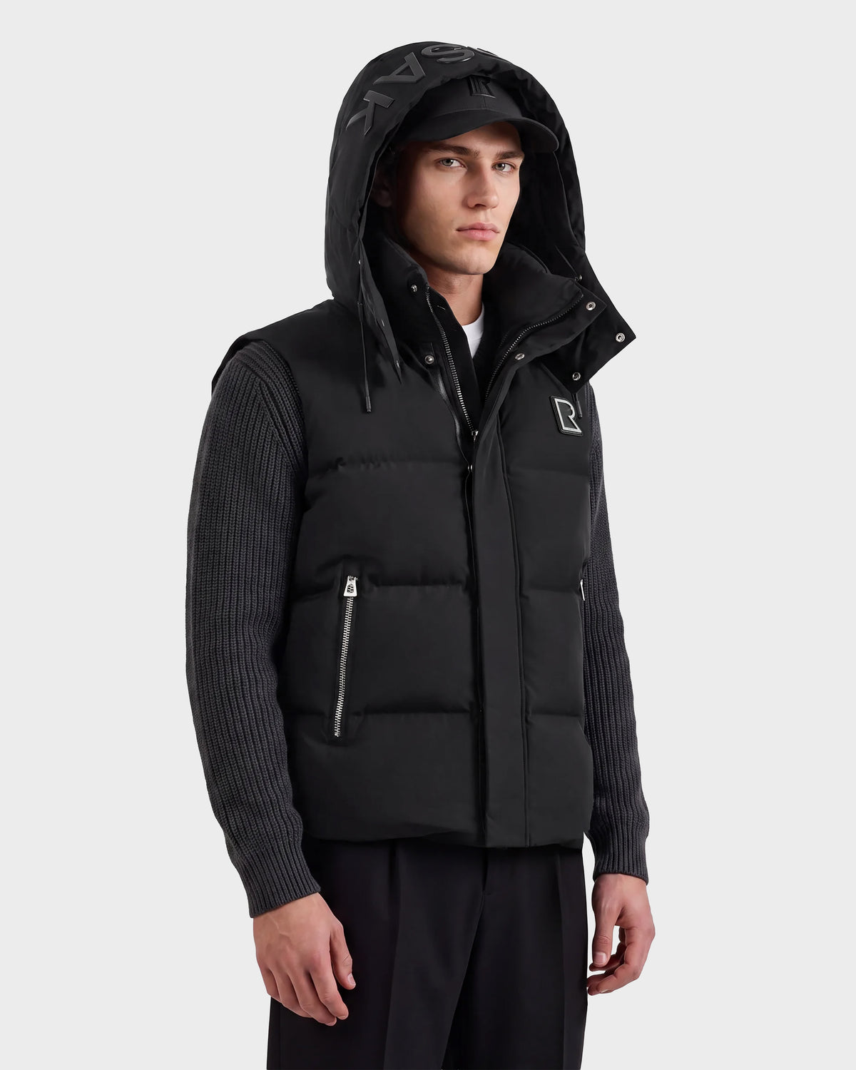 A man wearing the Rudsak REN M - 6125691 Black quilted vest with a hood over a ribbed dark sweater and black pants stands sideways, looking at the camera with a neutral expression against a plain light background.