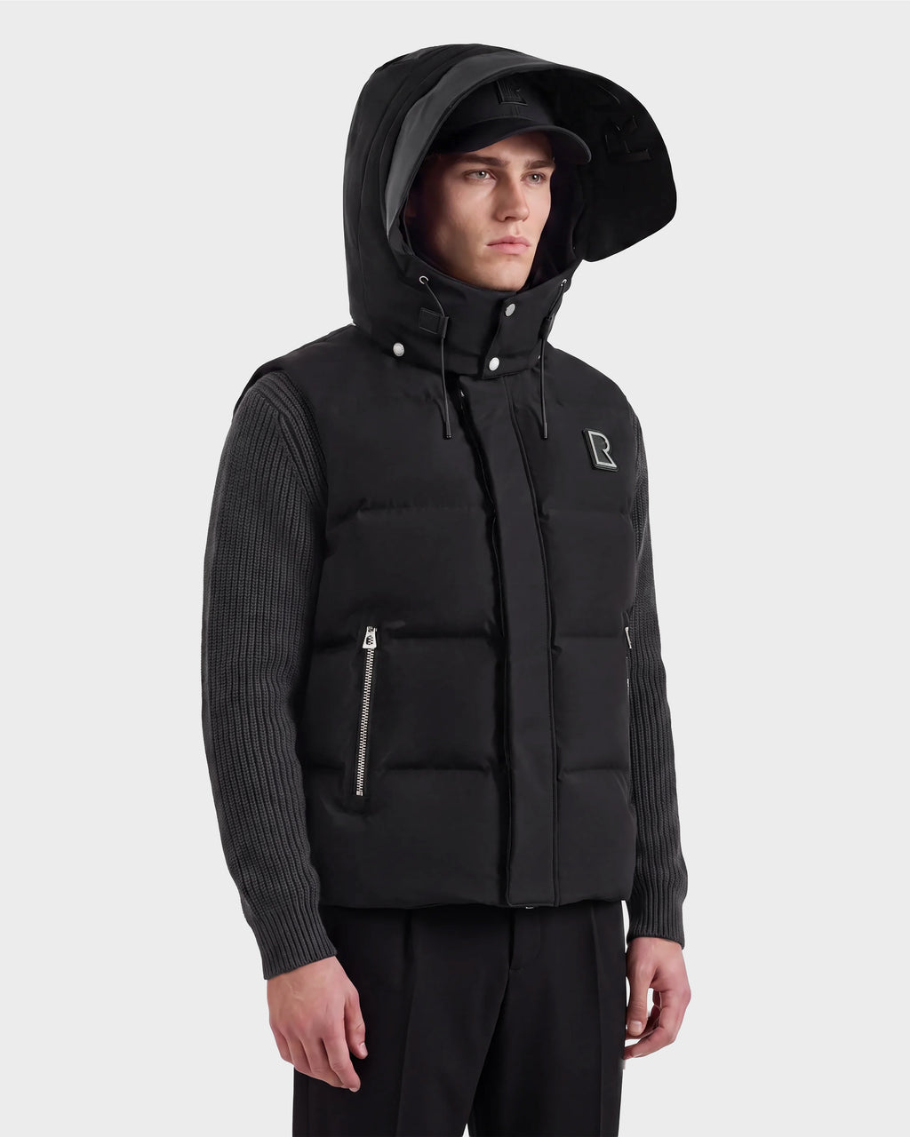 A man wearing the REN M - 6125691 Black down puffer vest with a hood from Rudsak over a ribbed long-sleeve shirt, paired with black pants and a cap, stands against a plain light background.