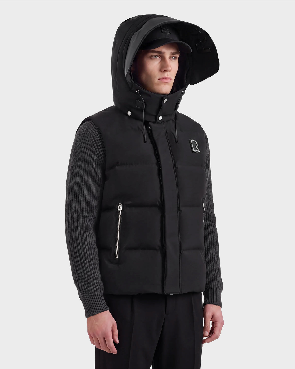 A man wearing the REN M - 6125691 Black down puffer vest with a hood from Rudsak over a ribbed long-sleeve shirt, paired with black pants and a cap, stands against a plain light background.