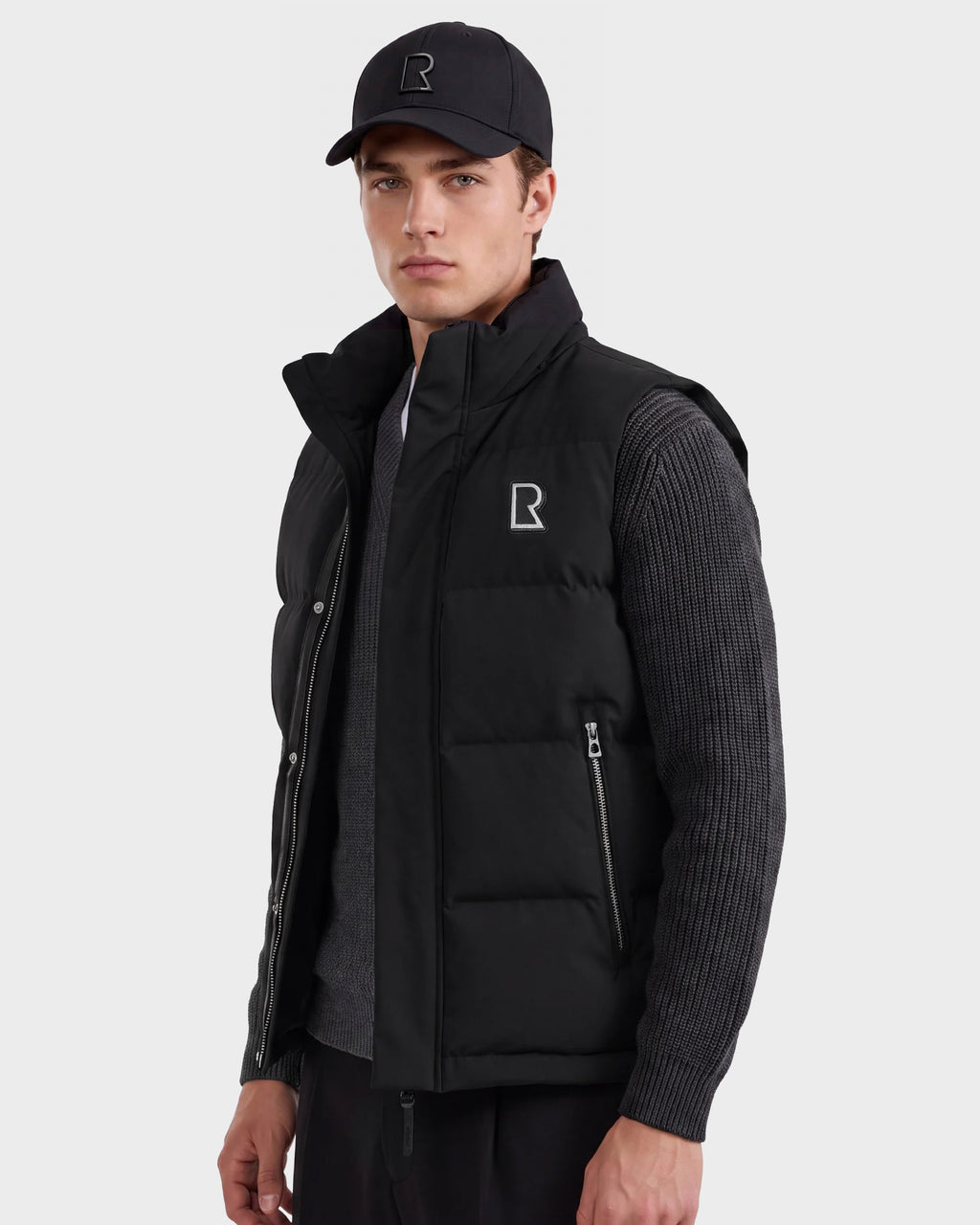 A man wearing a black cap, a Rudsak REN M - 6125691 Black men’s down puffer vest with the “R” logo, and a gray ribbed sweater stands facing forward against a plain background.