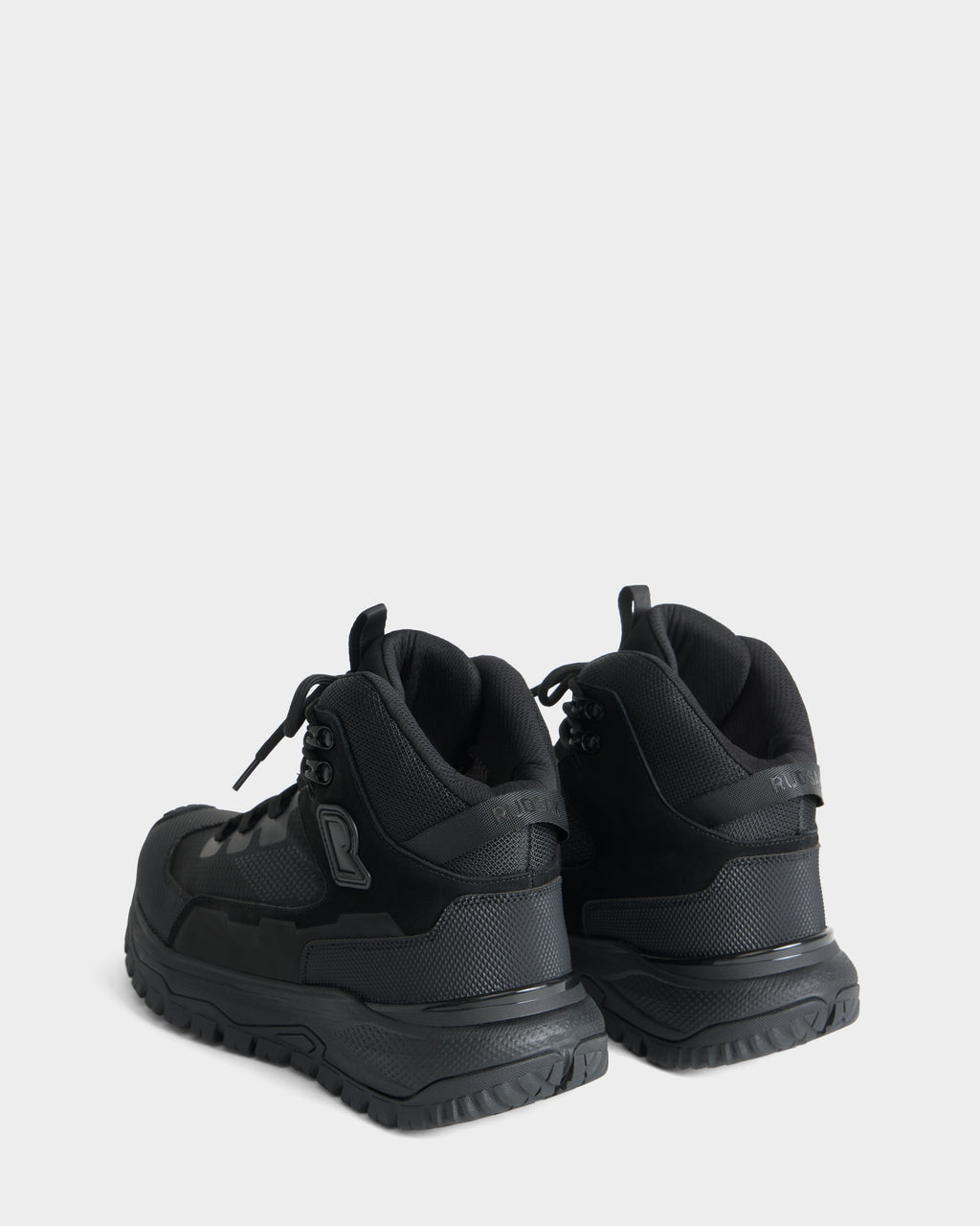 A pair of REXON - 6226503 Black high-top winter boots from Rudsak are displayed from a rear angle, highlighting their textured soles and padded collars against a plain light gray background.