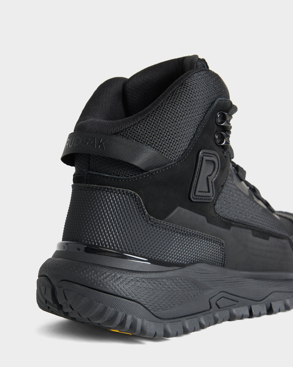 Close-up of the Rudsak REXON - 6226503 Black athletic high-top sneaker, highlighting its textured materials, thick treaded sole, and embossed R logo on the side. The heel and back part of the shoe are visible against a plain background.
