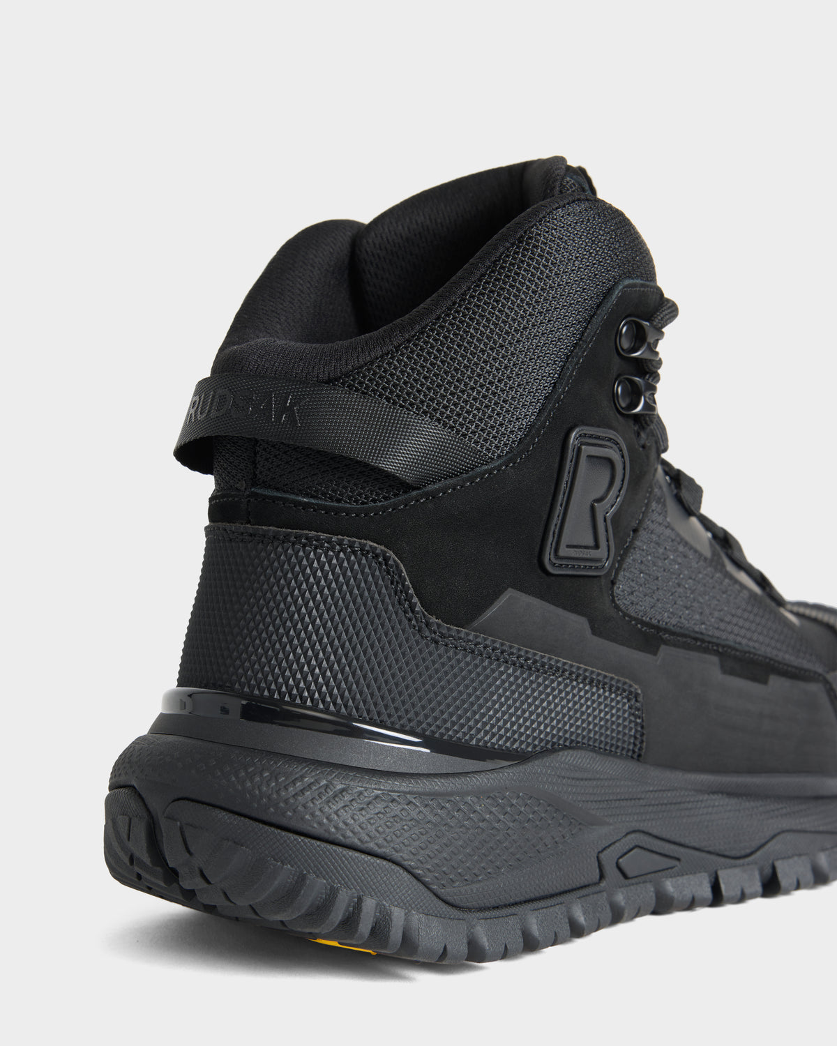 Close-up of the Rudsak REXON - 6226503 Black athletic high-top sneaker, highlighting its textured materials, thick treaded sole, and embossed R logo on the side. The heel and back part of the shoe are visible against a plain background.