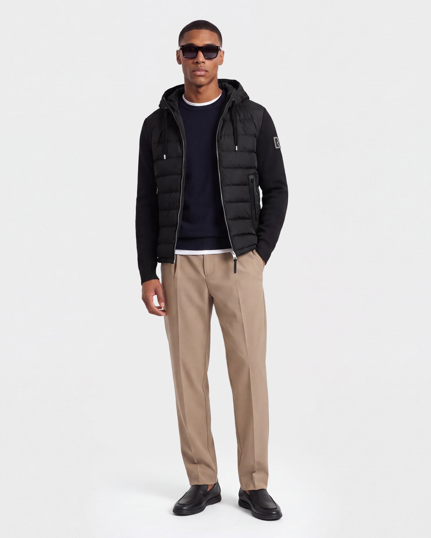 A man stands against a plain background wearing sunglasses, a black Rudsak RIVER - 6124584 down jacket, a dark sweater, tan dress pants, and black shoes.