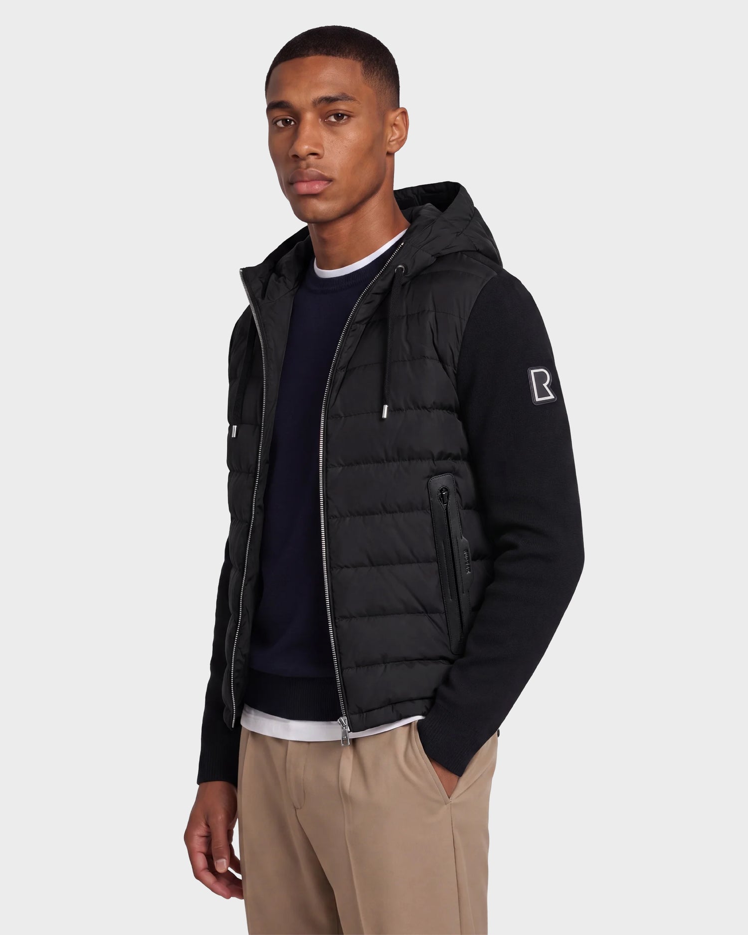 A man wearing the Rudsak RIVER - 6124584 Black hooded down jacket stands against a plain light background, paired with a navy shirt and tan pants. He faces the camera with a neutral expression.