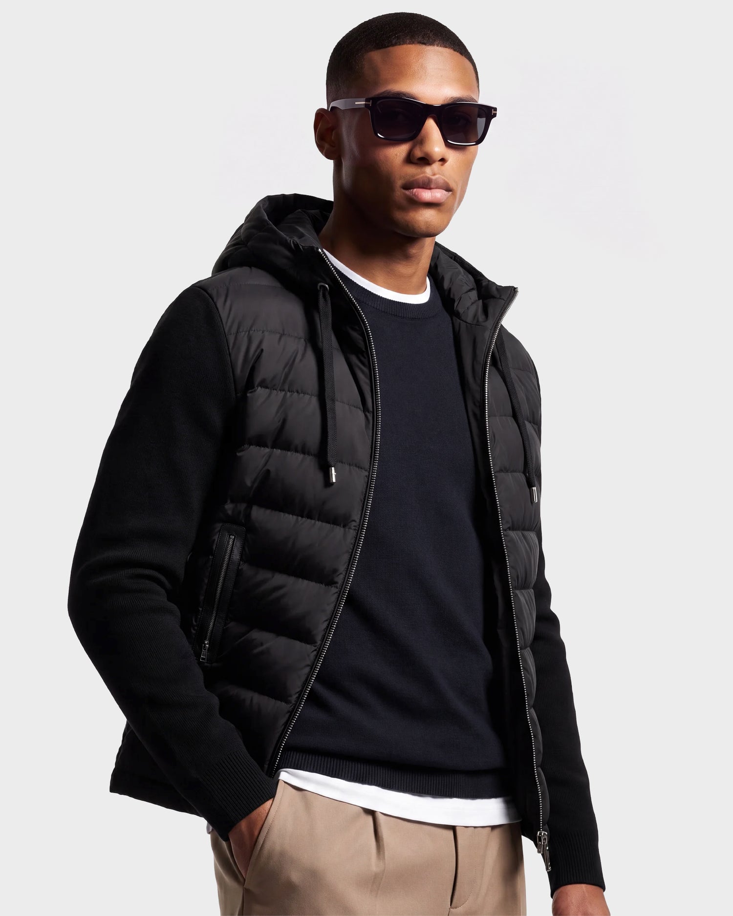 A man wearing sunglasses stands confidently against a plain light background, dressed in the Rudsak RIVER - 6124584 Black hybrid down jacket with a quilted hoodie over a dark sweater and white shirt, paired with beige pants.