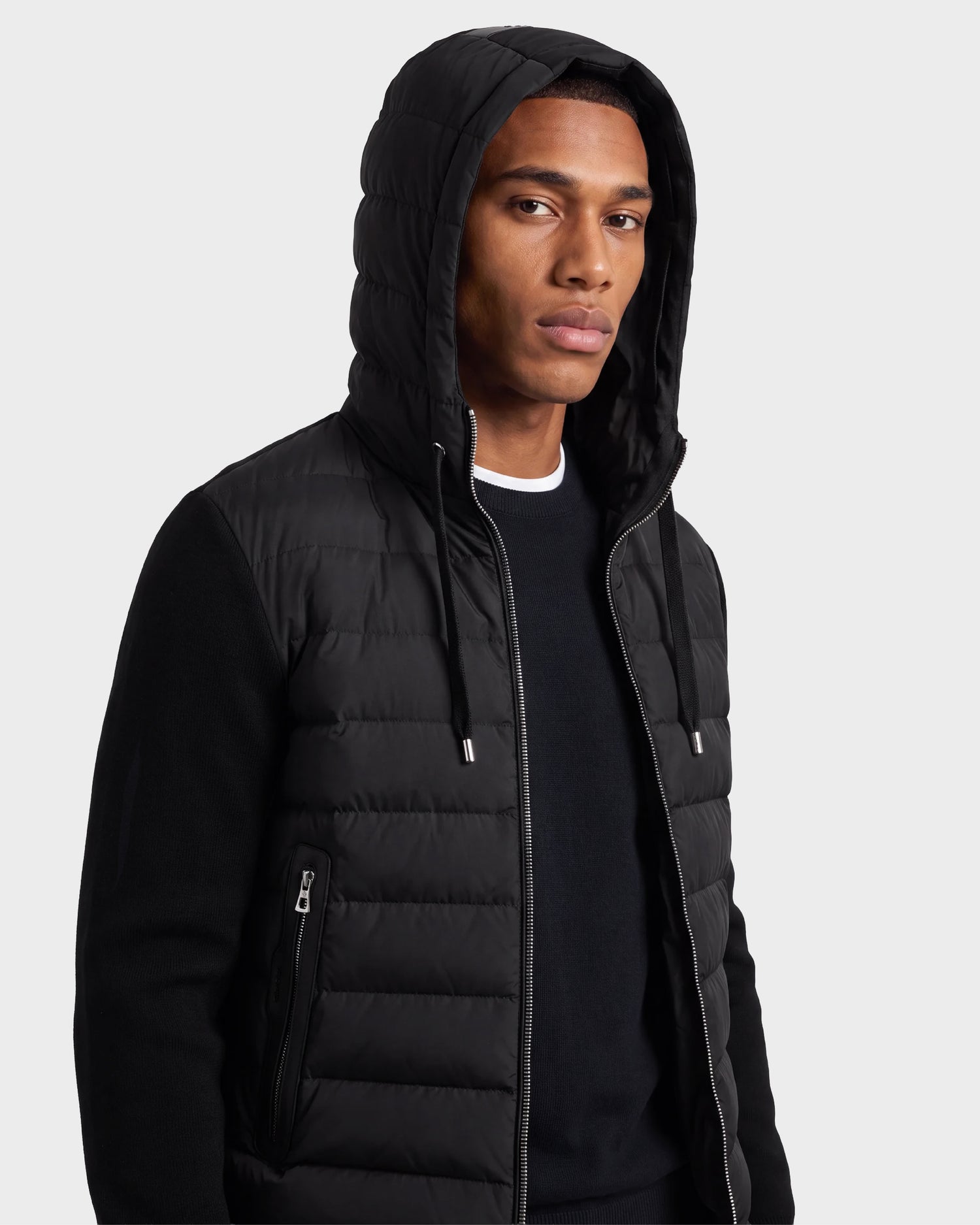 A man wearing the Rudsak RIVER - 6124584 Black hooded down jacket over a black sweater stands against a plain light background, looking slightly to the side with a neutral expression.