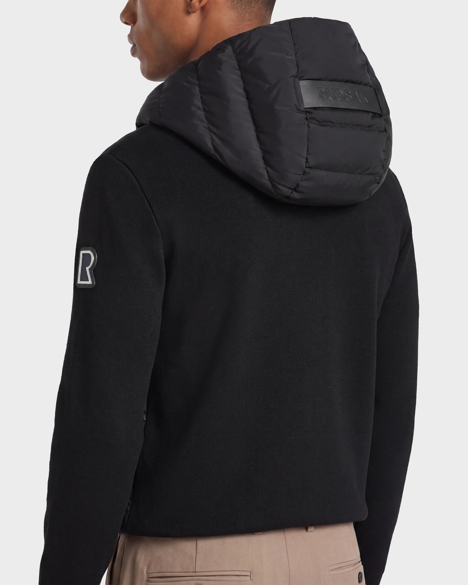 A man is seen from behind wearing the Rudsak RIVER - 6124584 black hybrid down jacket with a quilted hood, featuring a BOSS patch on the back and the letter "R" on the upper right sleeve, paired with beige pants.
